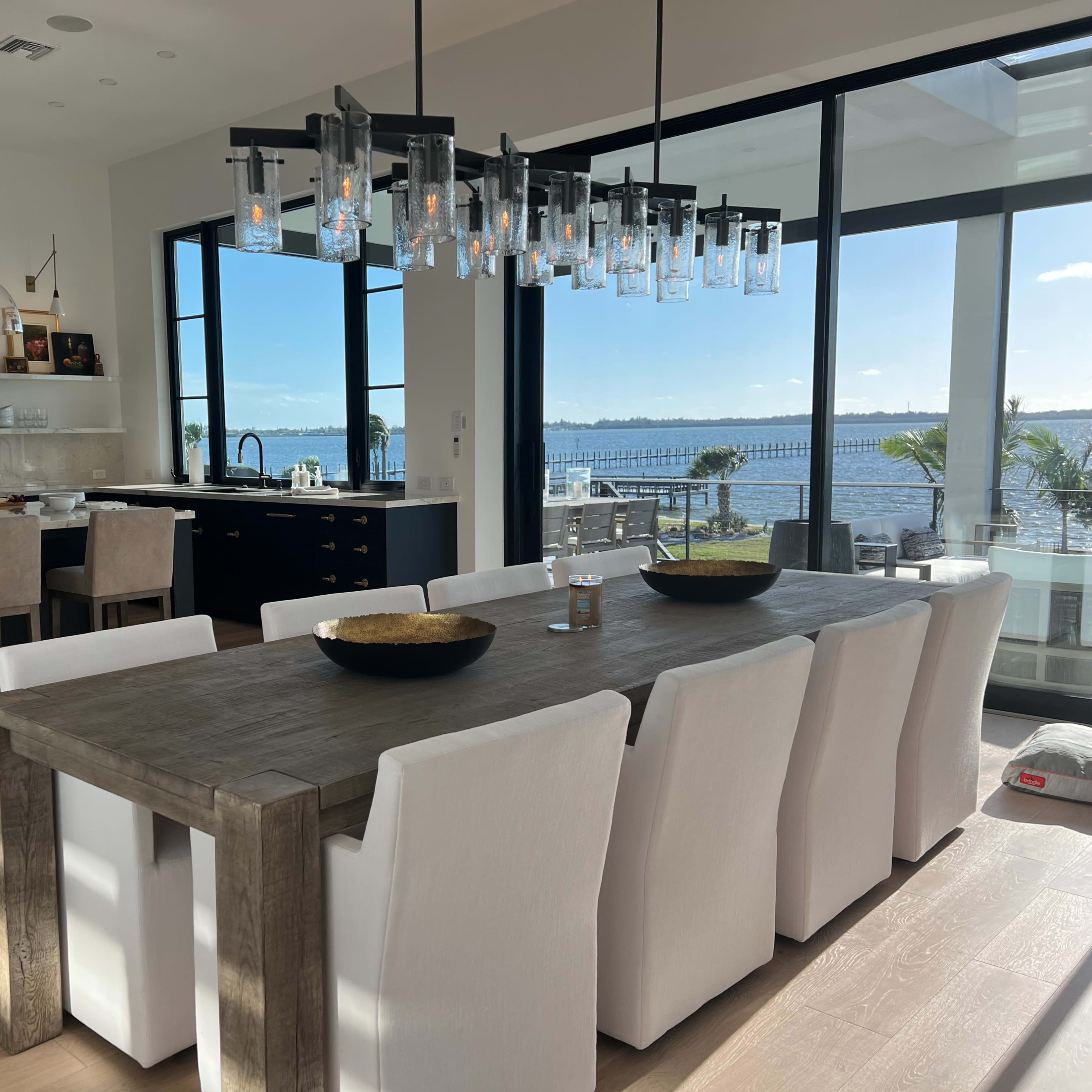 A modern dining area features a long wooden table surrounded by white upholstered chairs, with large windows showcasing a view of the water.