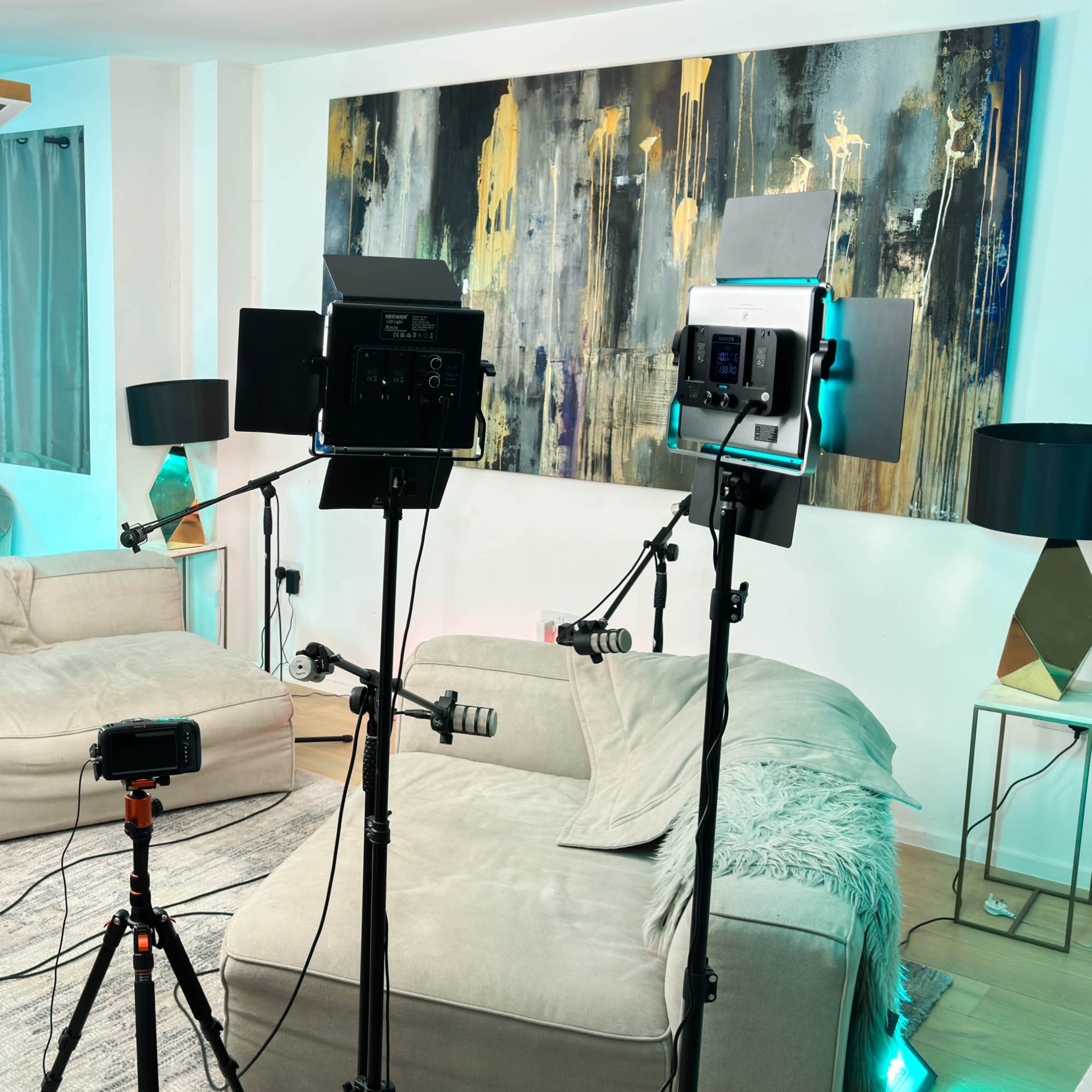 A well-lit studio setup in a living room, featuring two sofas, multiple filming lights, cameras on tripods, and a control panel on the floor.