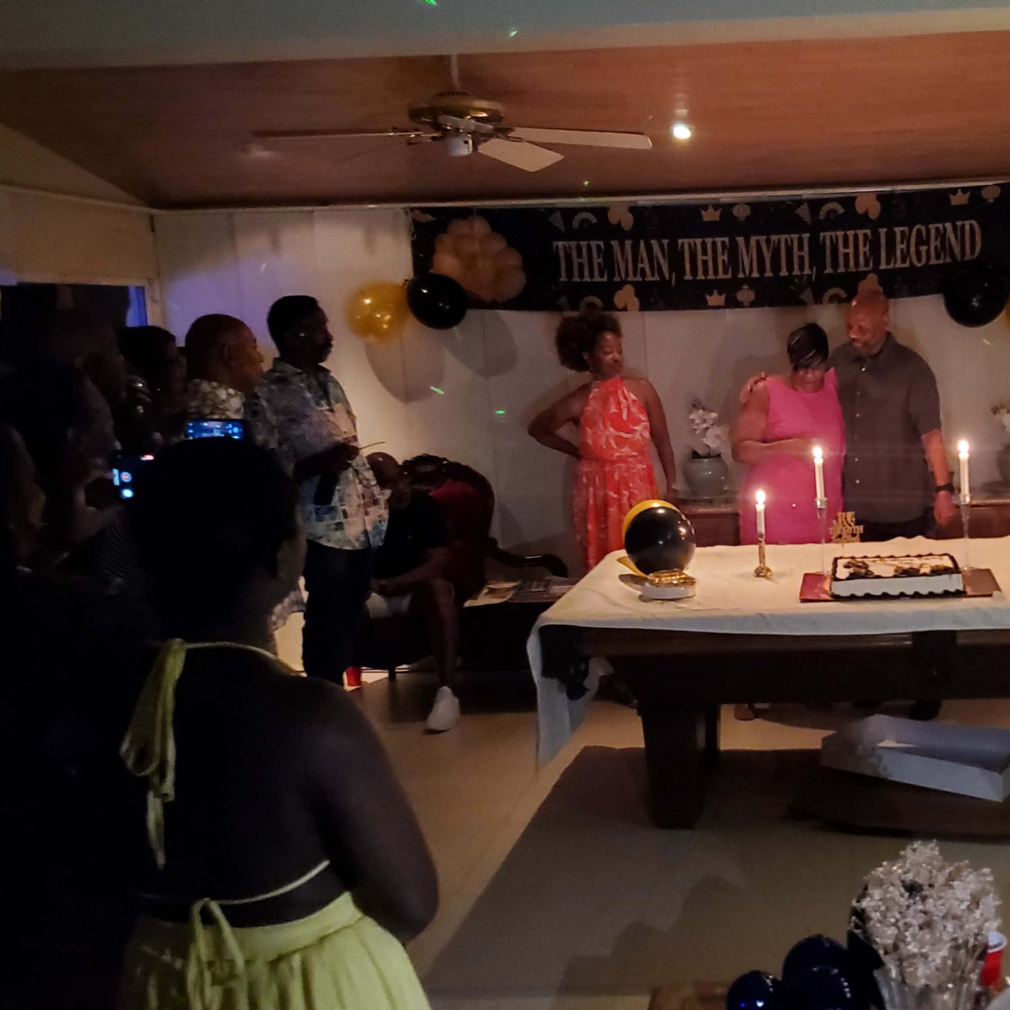 A group of people gathers around a table with a decorated cake and candles during a celebration in a well-lit living room.