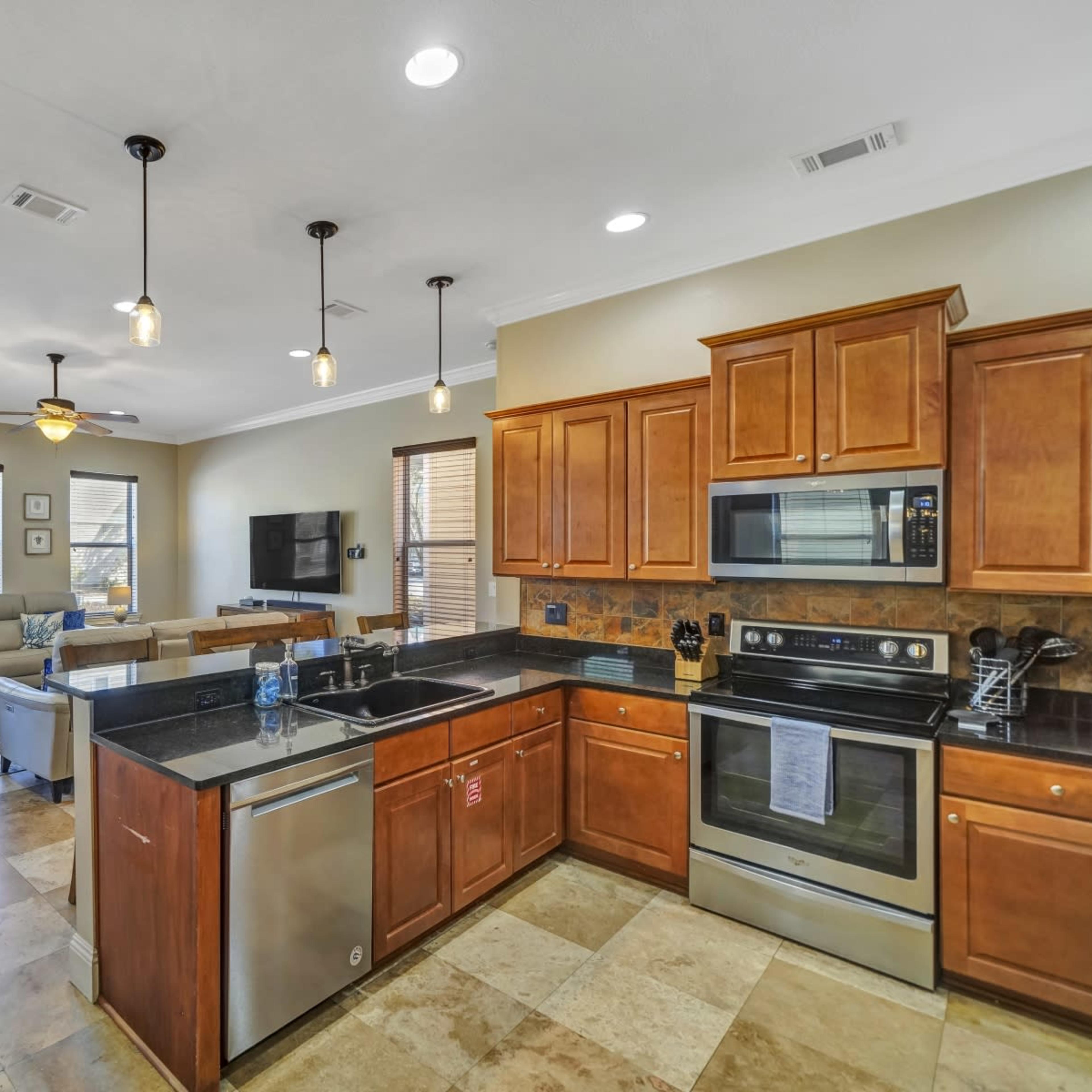 A modern kitchen with wooden cabinets and stainless steel appliances, opening into a living area with sofas and a television.