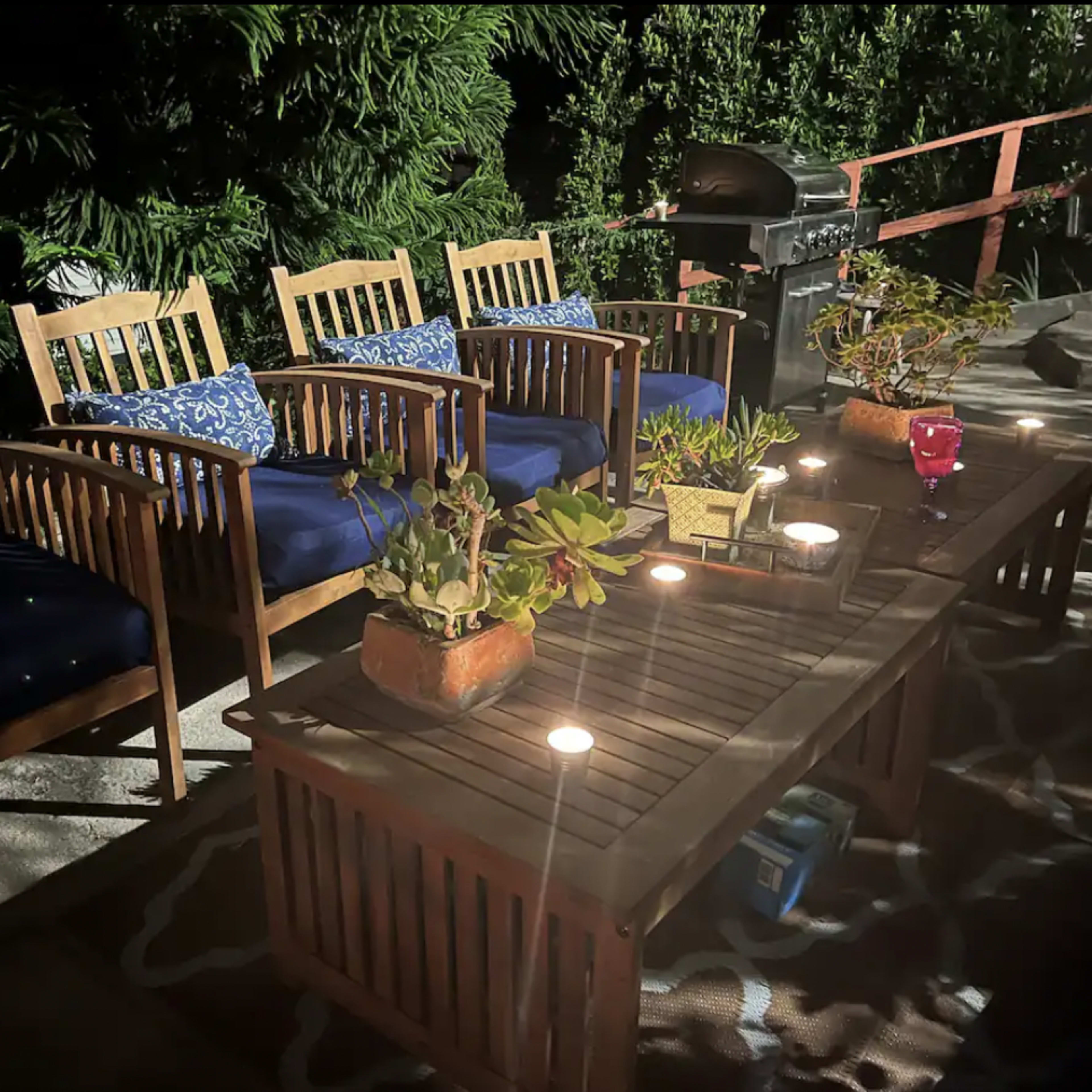 A set of four wooden chairs with blue cushions is arranged around a wooden coffee table with candles and potted plants, illuminated by nighttime lighting in a garden setting.