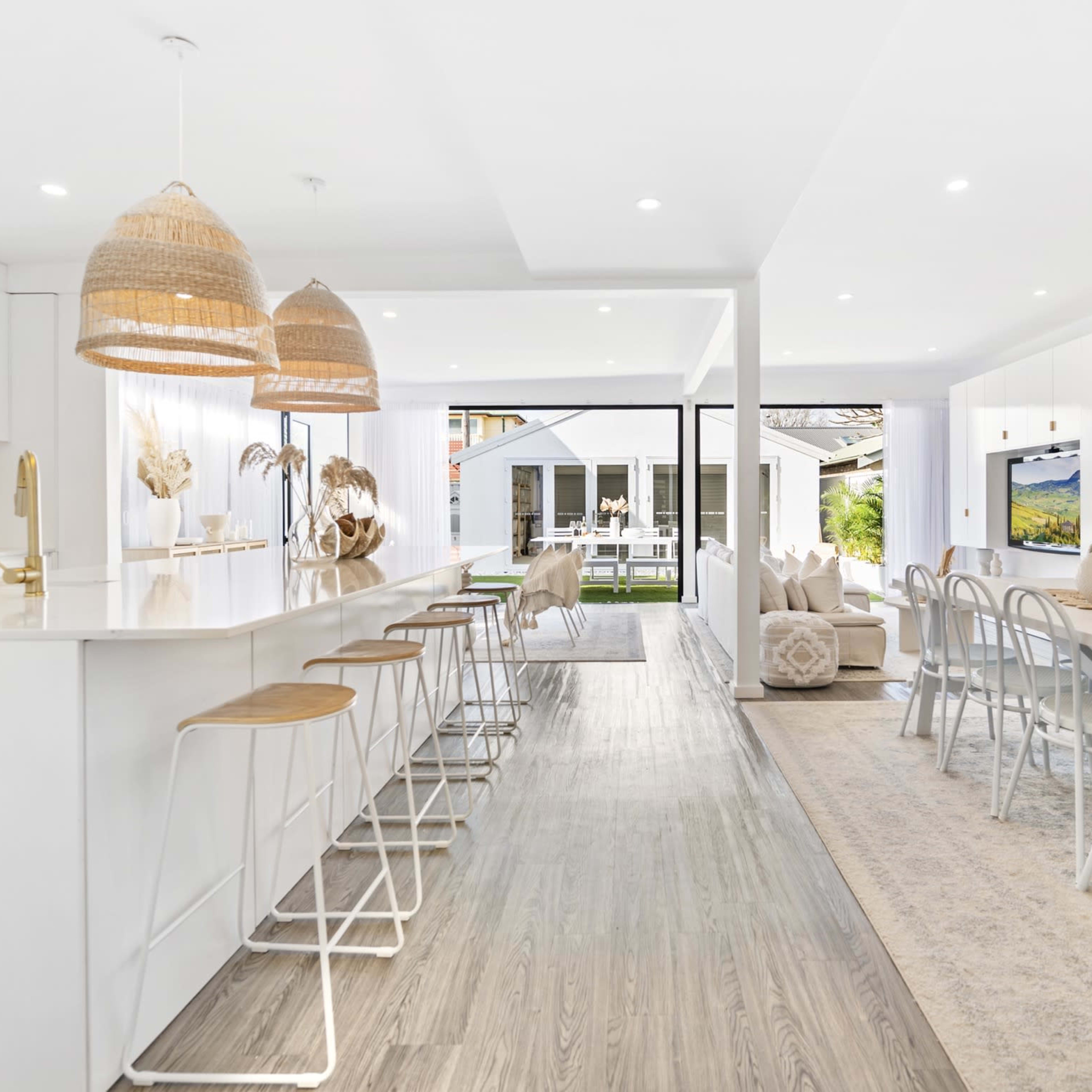 A modern, open-concept kitchen and dining area with white cabinetry, bar stools, and large windows overlooking an outdoor space.