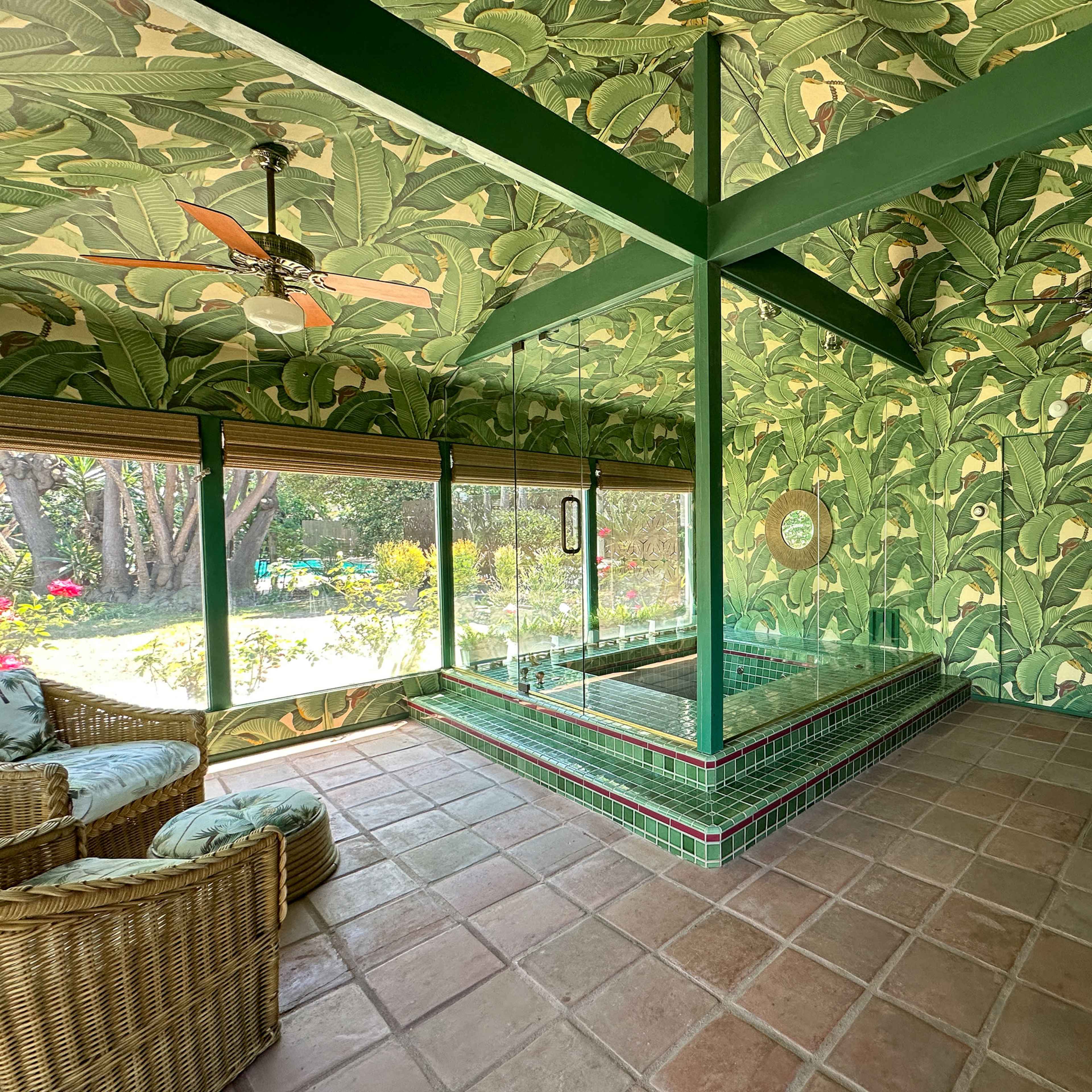 The spacious room features green tropical leaf-patterned wallpaper, a tiled jacuzzi, and wooden furniture.