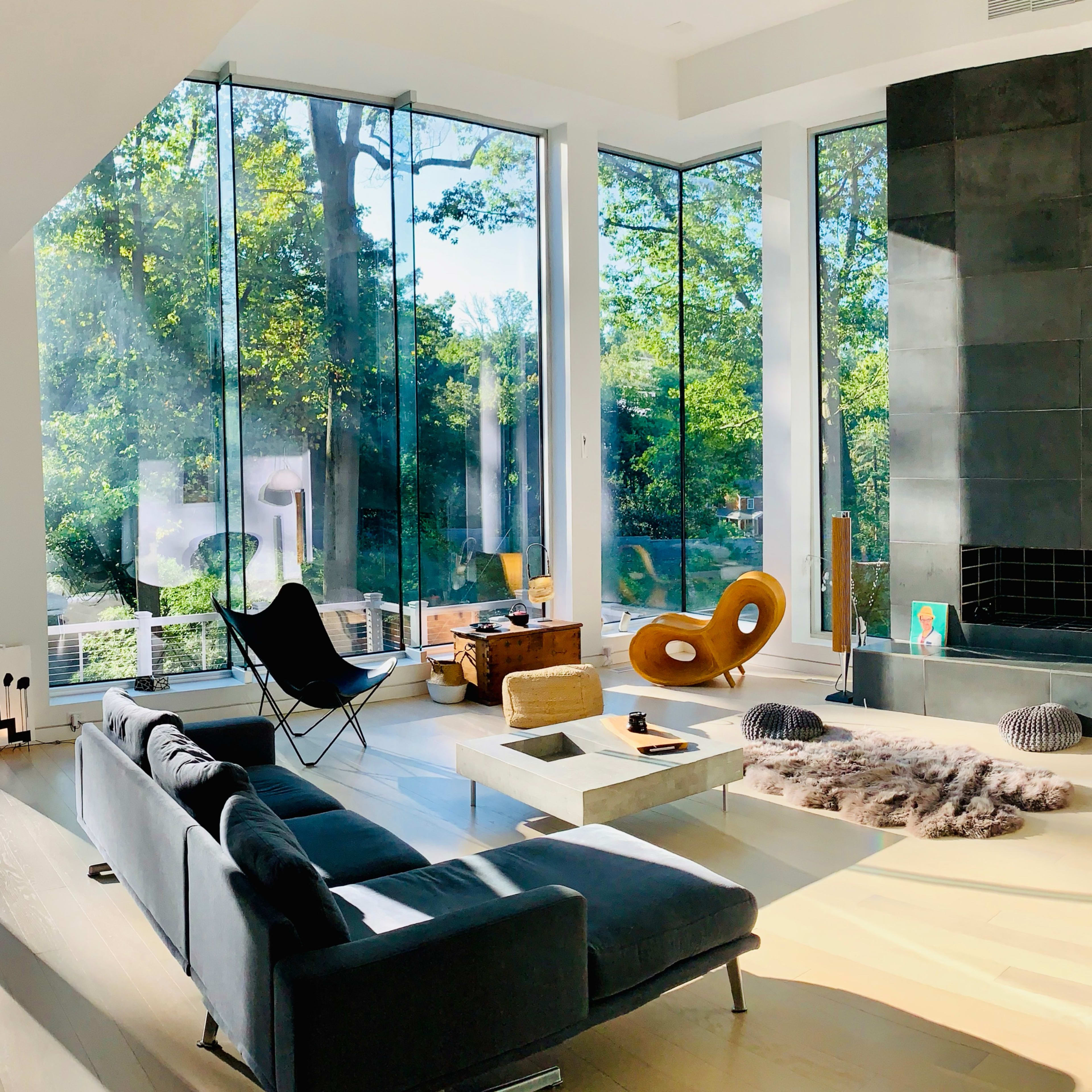 A modern living room features large glass windows, a stone fireplace, and contemporary furniture, overlooking a lush green landscape.