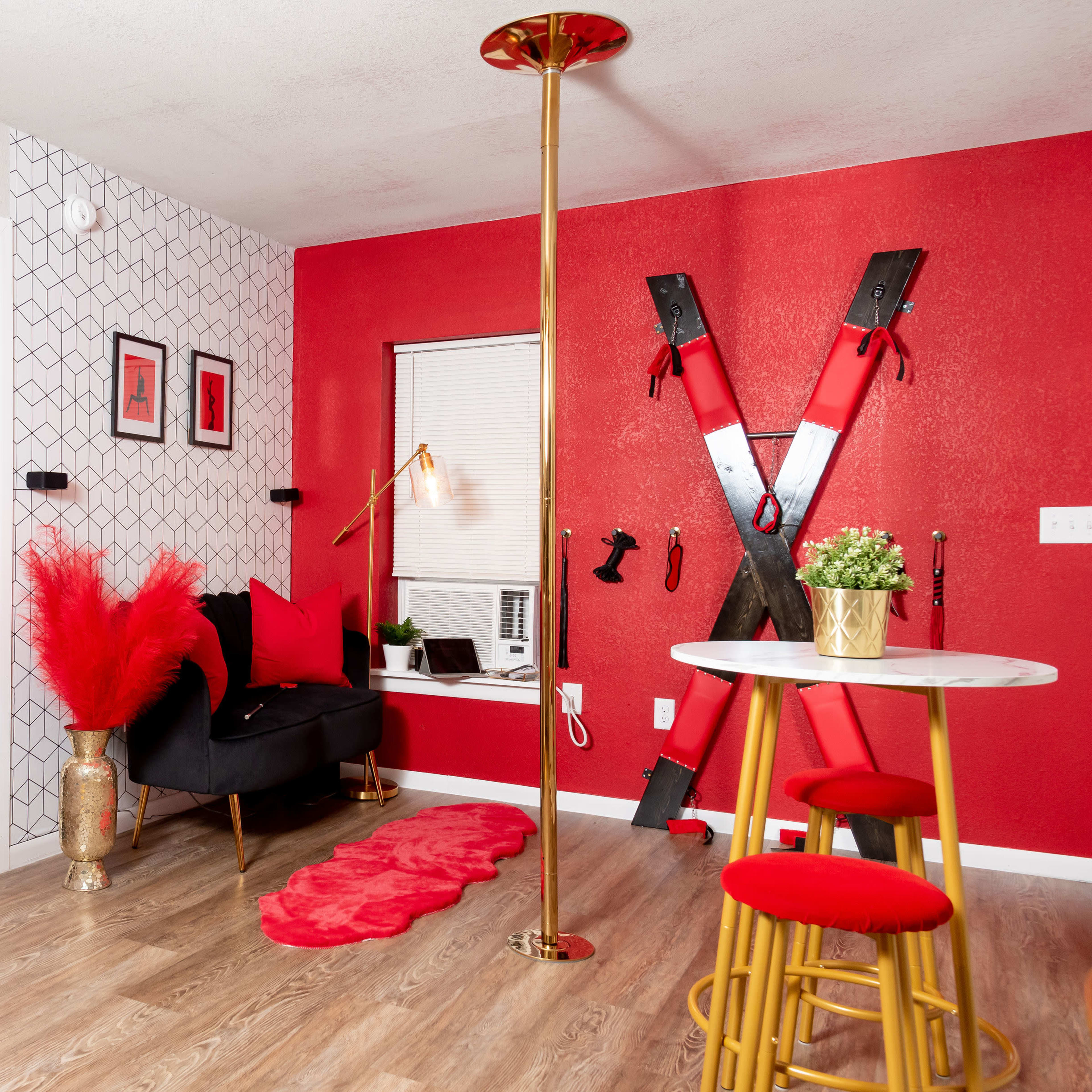 A small, modern kitchen and living area with a red accent wall, a black sofa, a dining table with yellow stools, and a decorative pole.