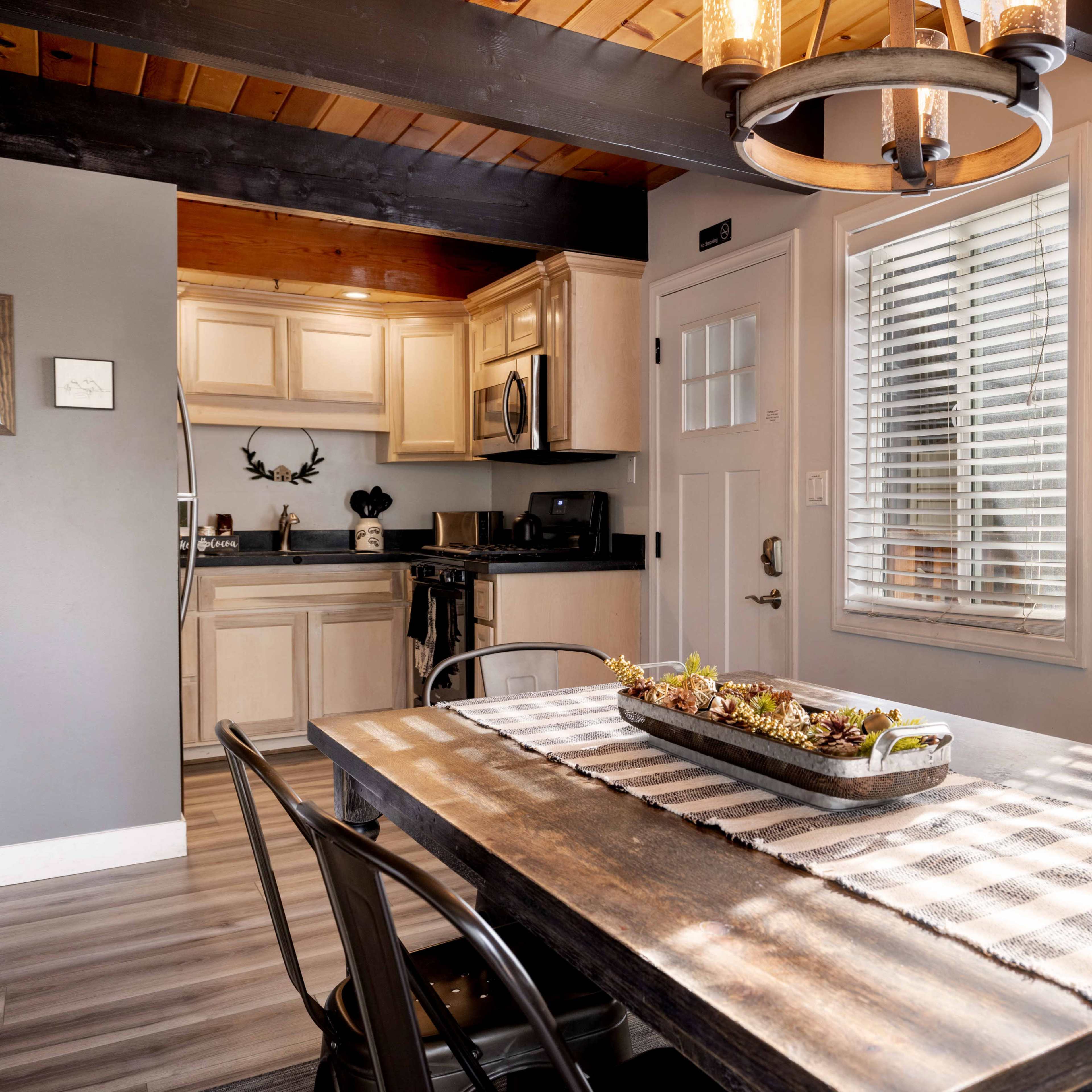 A dining area with a wooden table set with a decorative planter, surrounded by black chairs, leading to a kitchen with beige cabinetry visible in the background.
