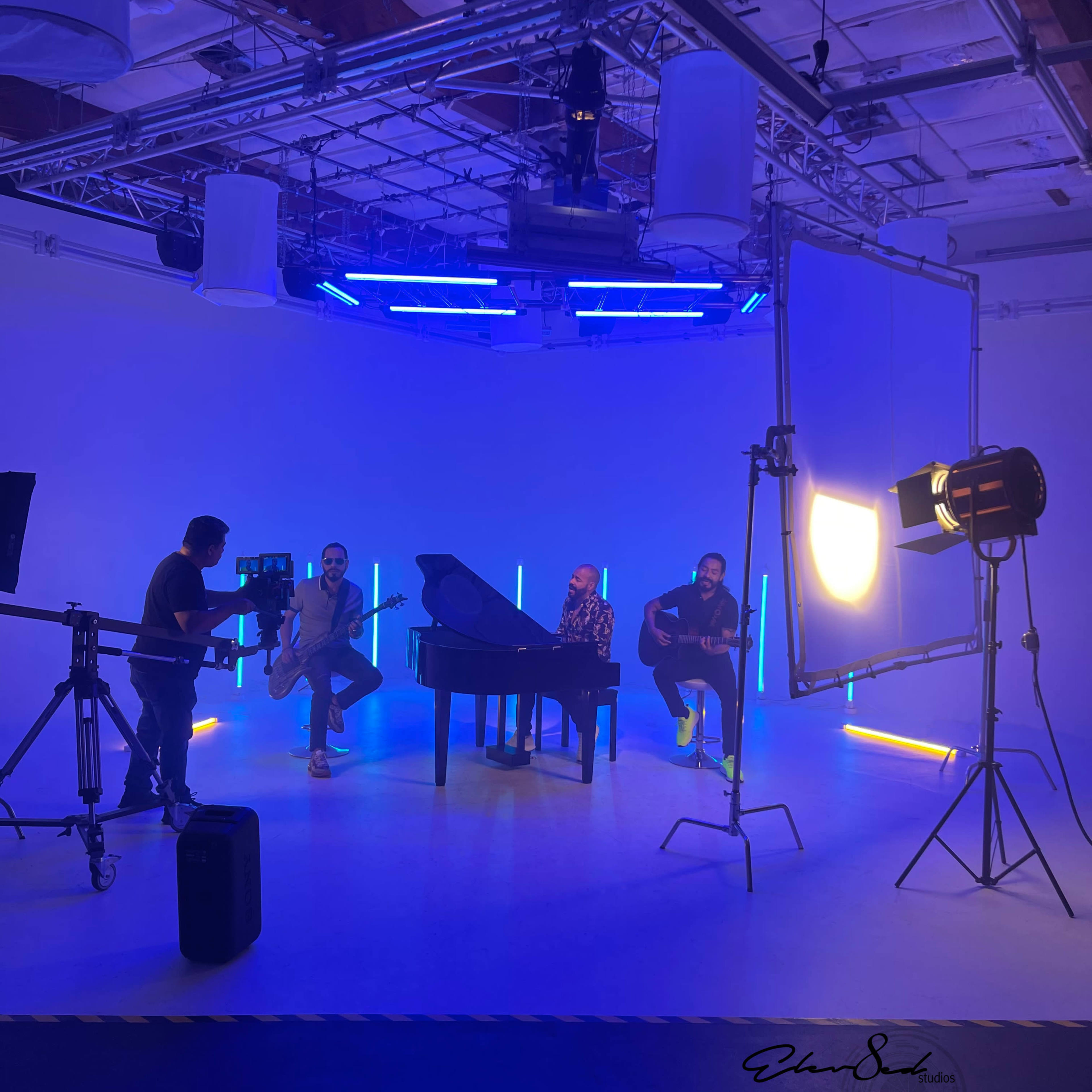 Four musicians perform in a studio setting with a grand piano and colorful lighting.