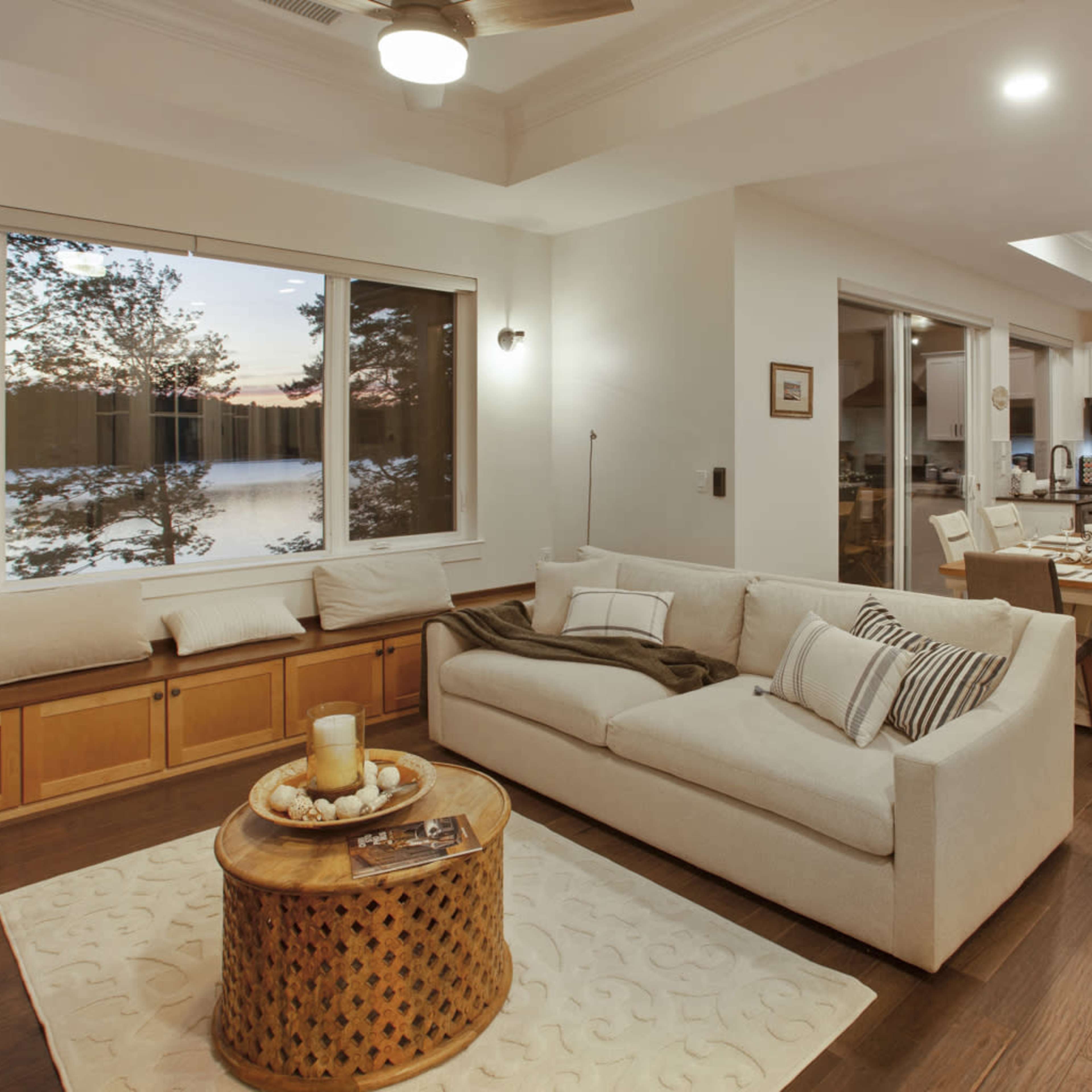 A modern living space featuring a beige sofa, a round wooden coffee table, a dining area, and large windows with a view of a lake at sunset.