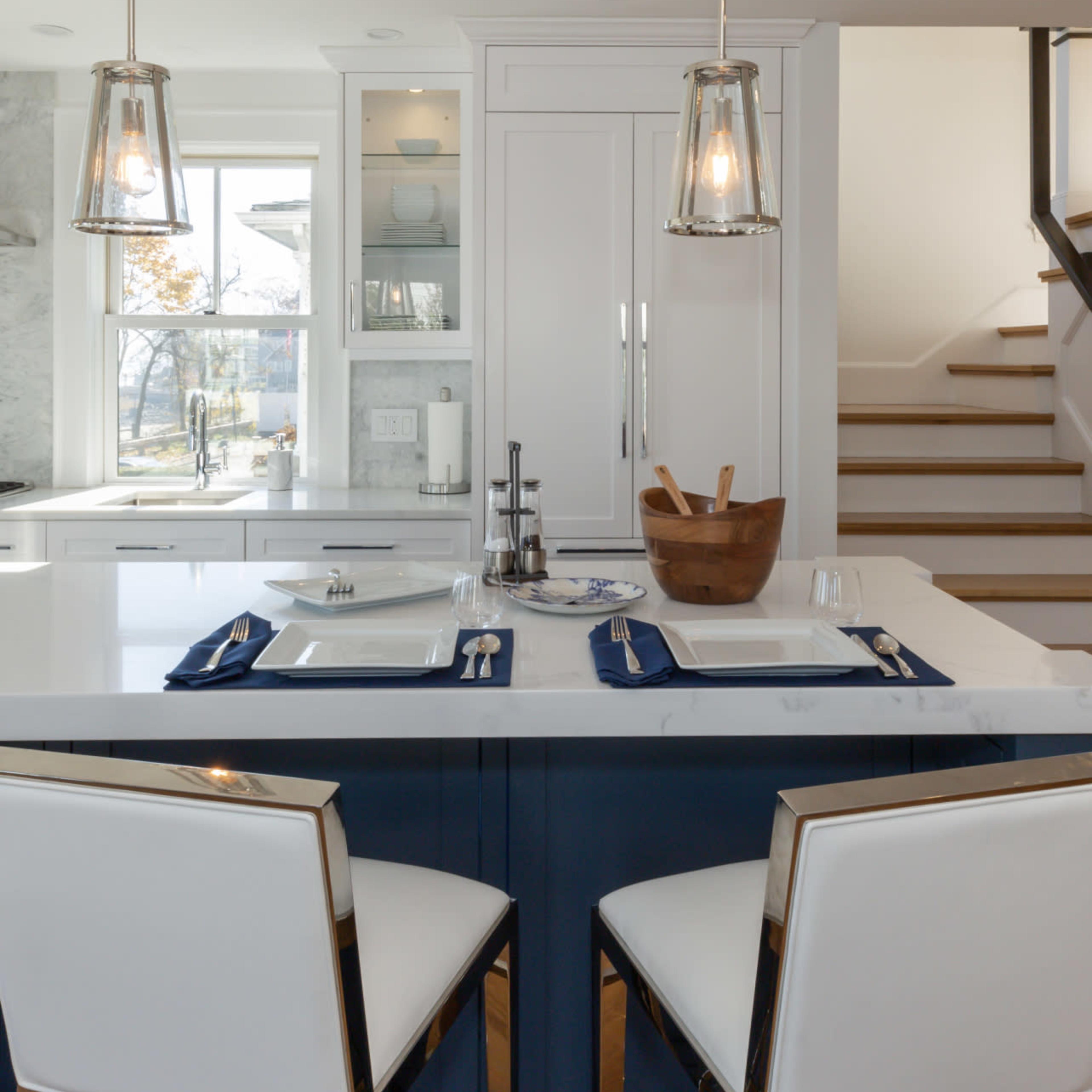 A modern kitchen features a white island with two bar stools, pendant lights overhead, and a neatly arranged dining area.