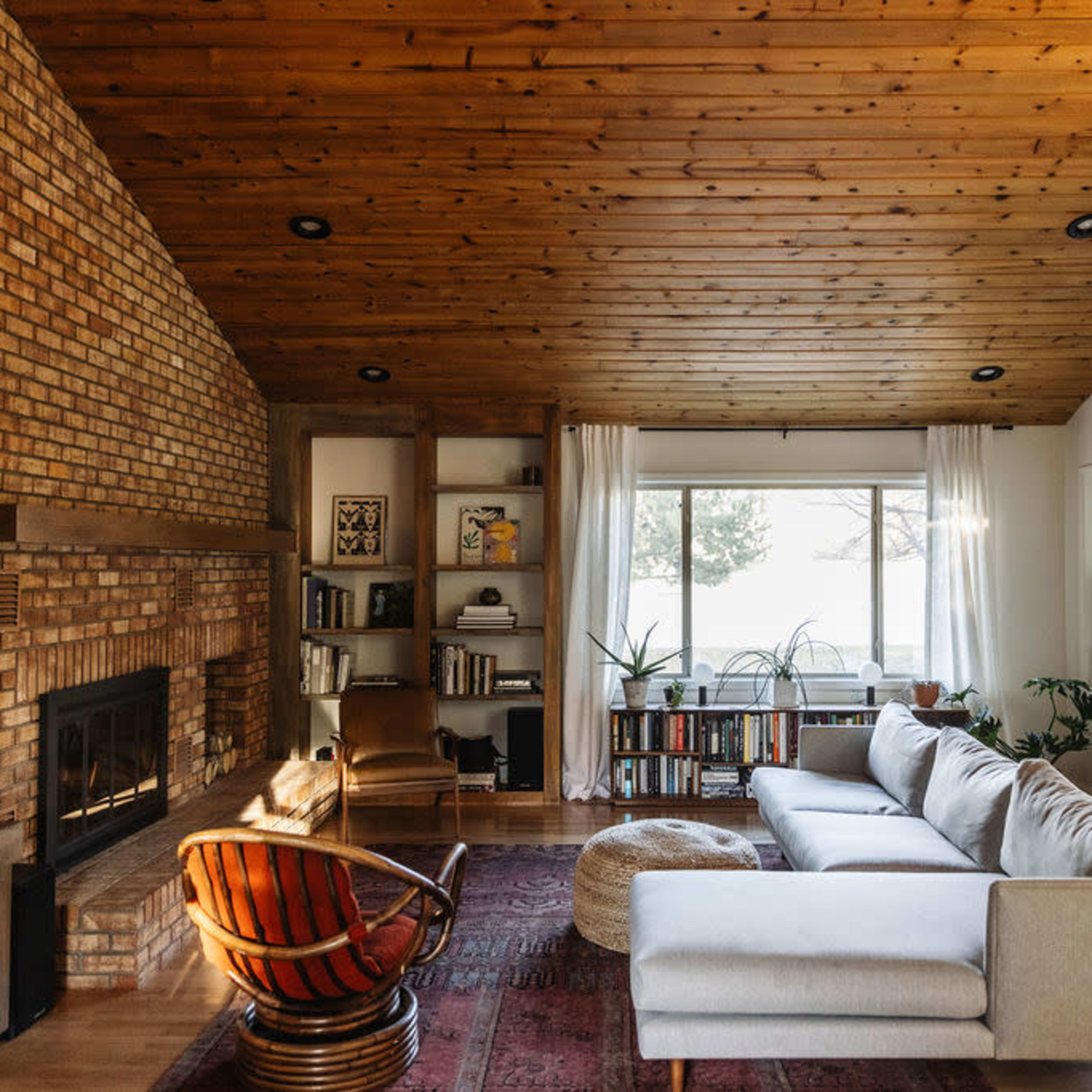 A cozy living room features a brick fireplace, wooden ceiling, large windows, and a sectional sofa with a circular ottoman.