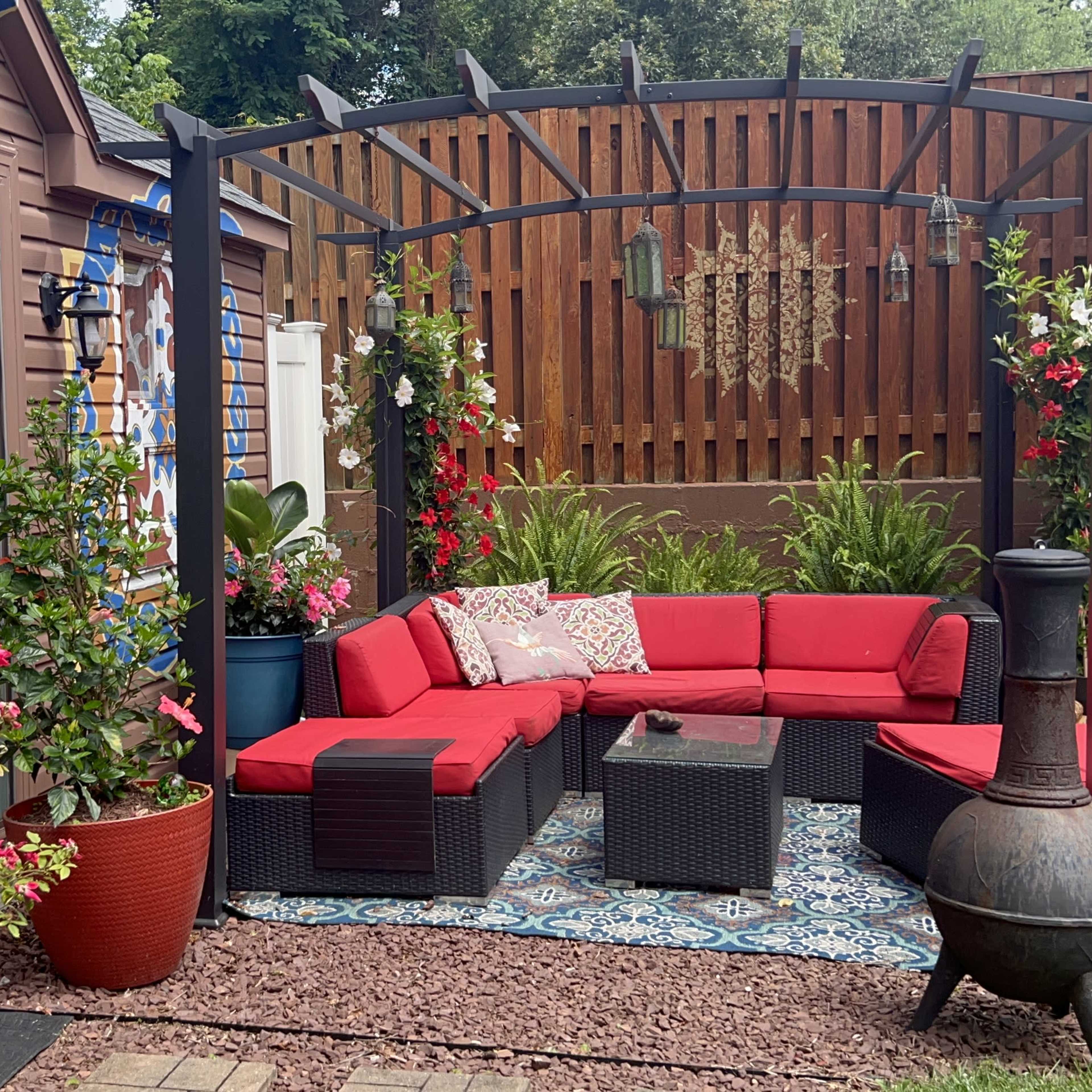 A cozy outdoor seating area with a red sectional sofa, a coffee table, decorative plants, and a barbecue grill in a landscaped yard.
