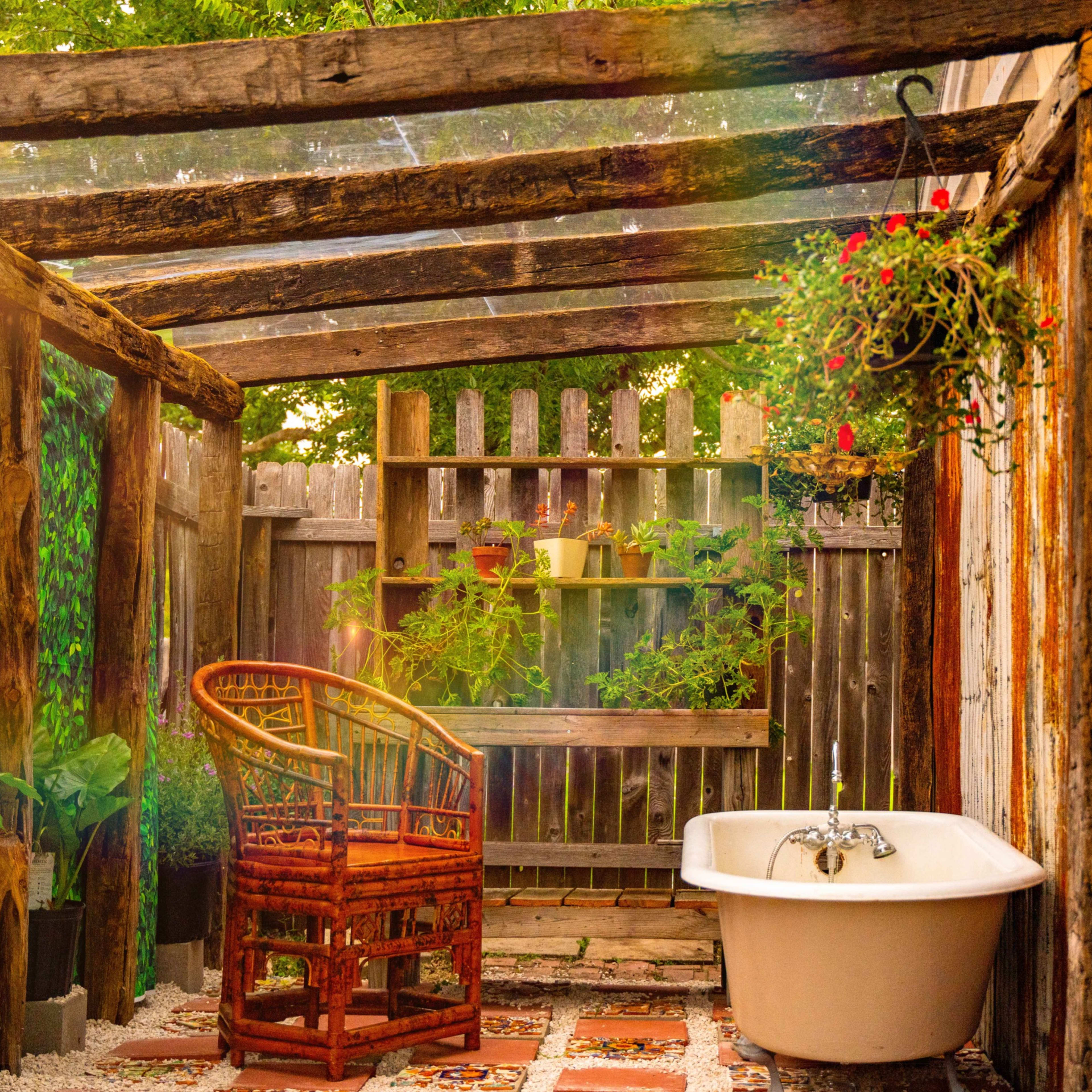 An outdoor bathroom space with a freestanding bathtub, a wicker chair, and potted plants surrounded by wooden structures and a transparent roof.