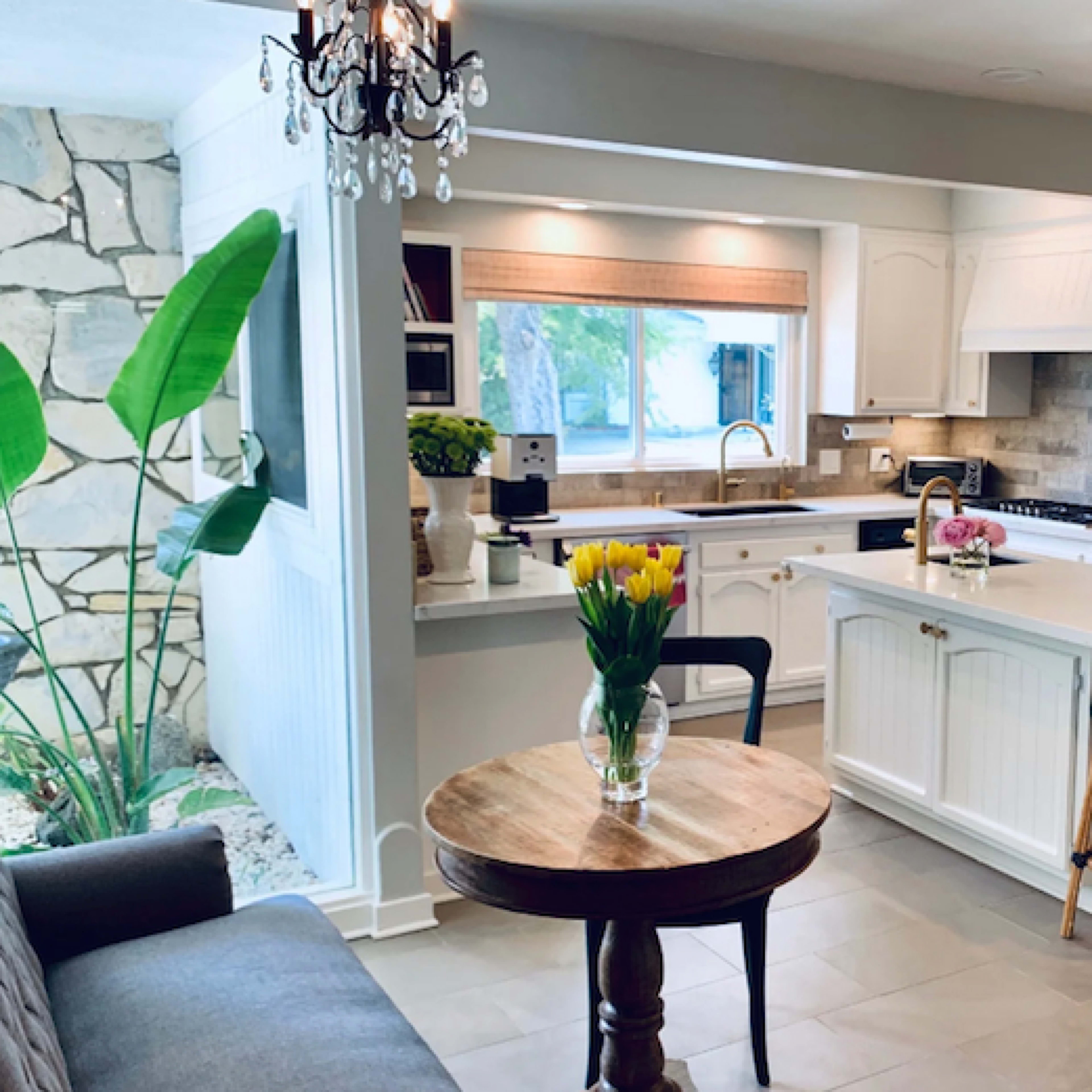 A modern kitchen features a central island with bar stools, white cabinetry, and a dining table with a vase of tulips, alongside large windows and tropical greenery visible outside.