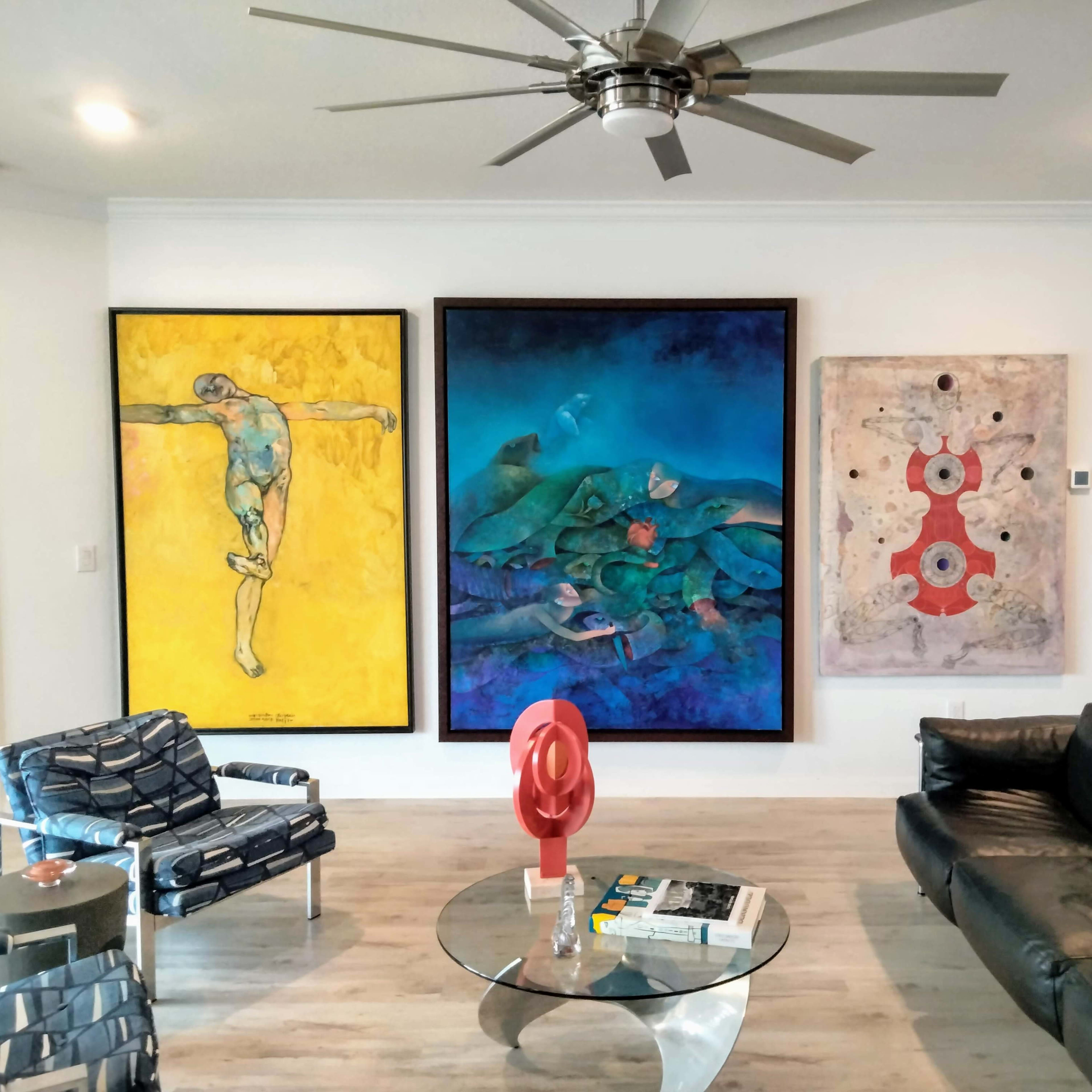 A living room with a black leather sofa, a glass coffee table, and three large colorful paintings mounted on the white wall.