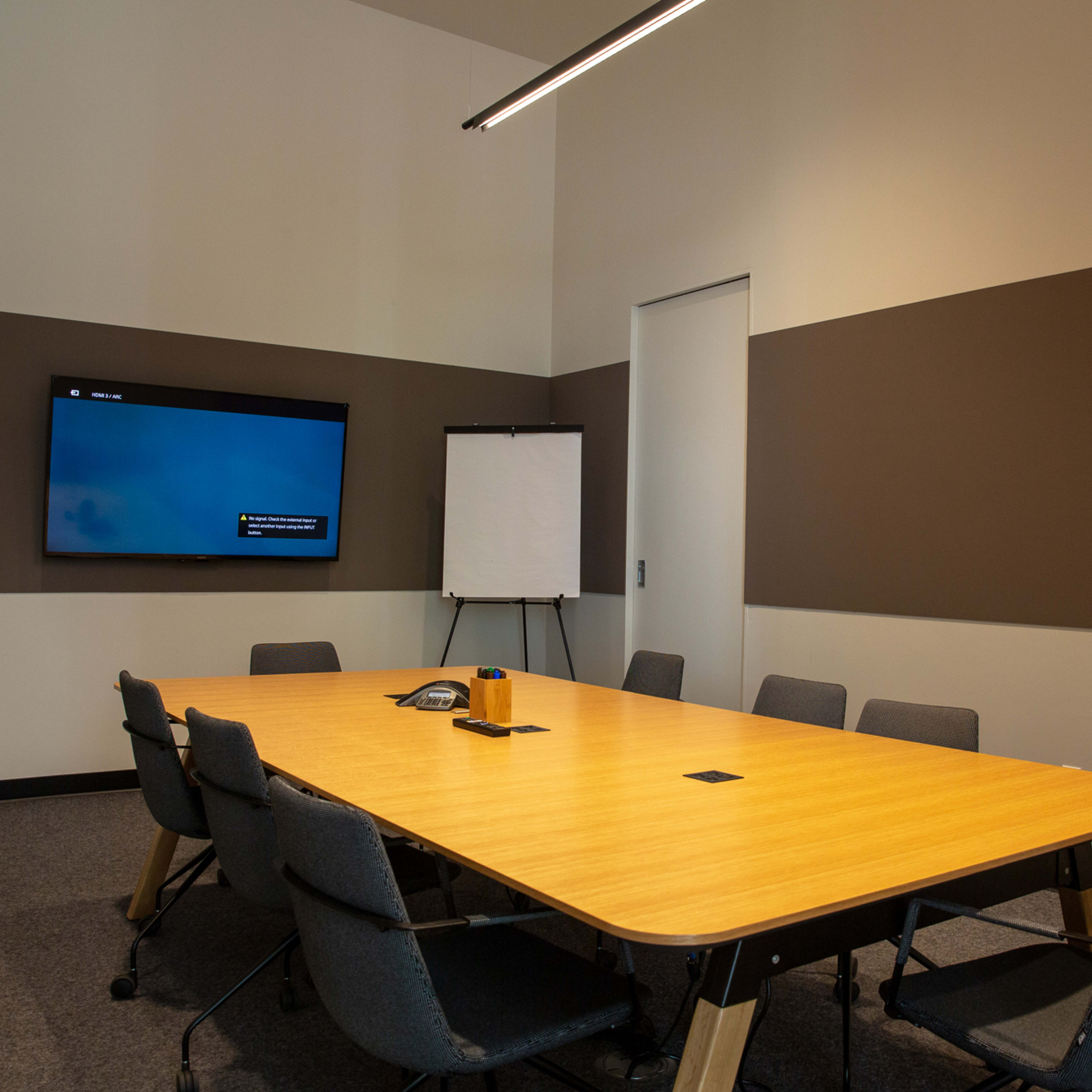 A conference room features a large wooden table surrounded by chairs, with a screen mounted on the wall and a flip chart nearby.