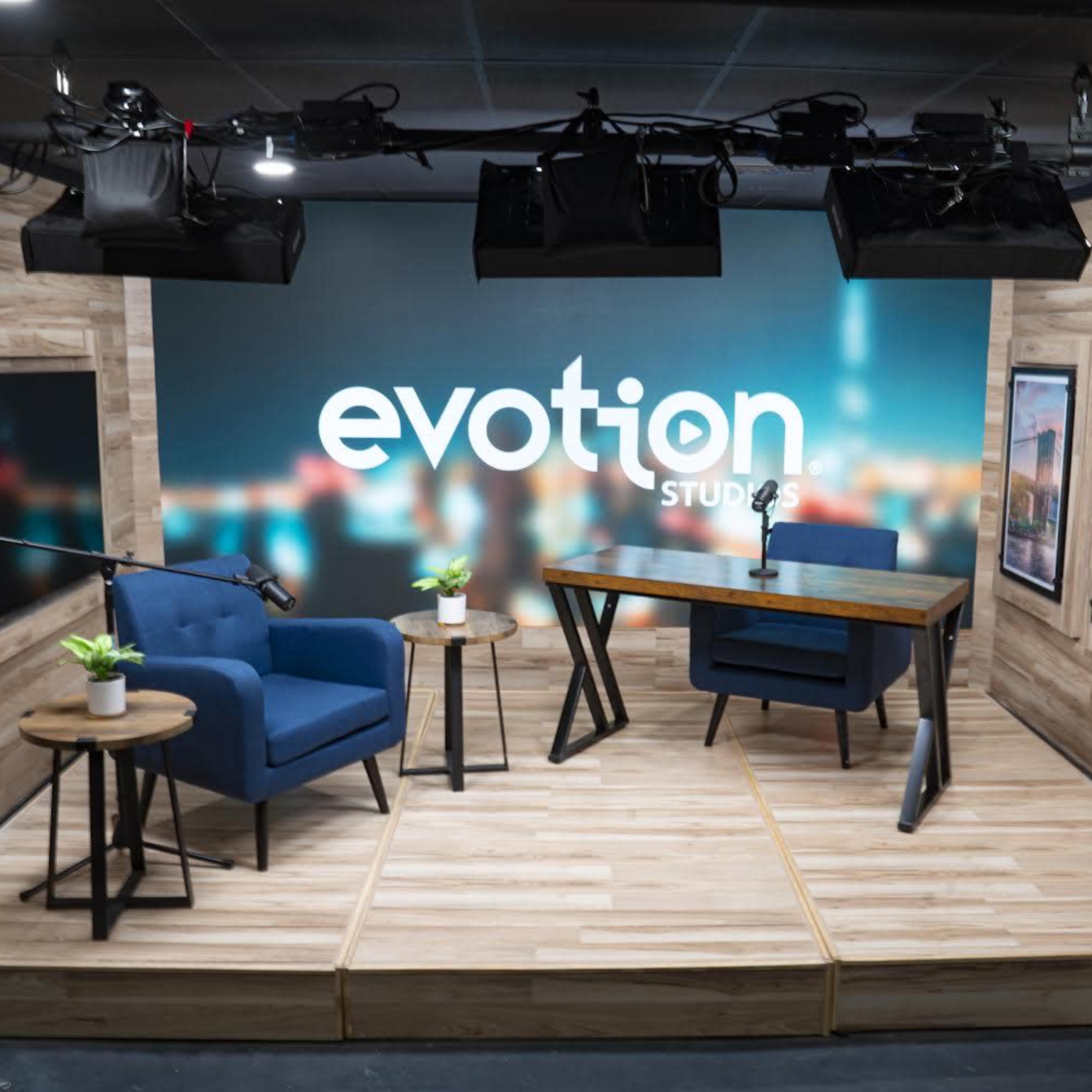 A modern studio setup features two blue chairs, a wooden table, and framed cityscape art on a wall with a large "evotion studios" logo.