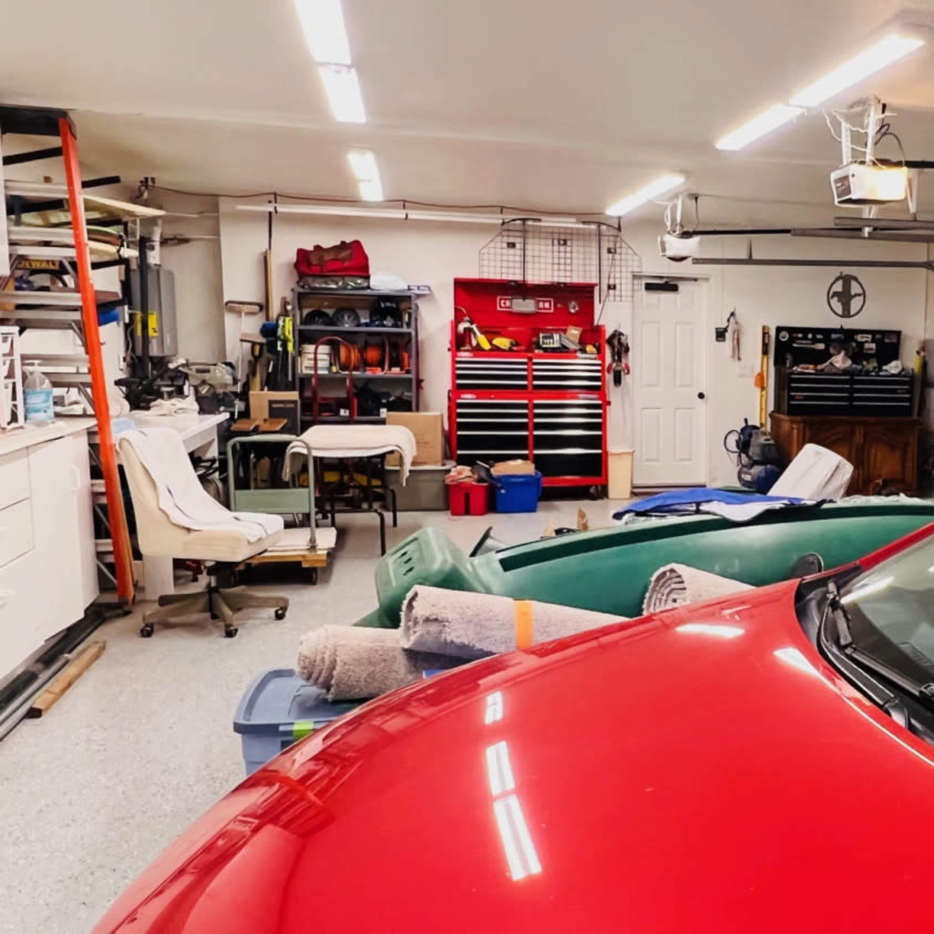 A red car is parked in a cluttered garage filled with various tools, equipment, and storage units.
