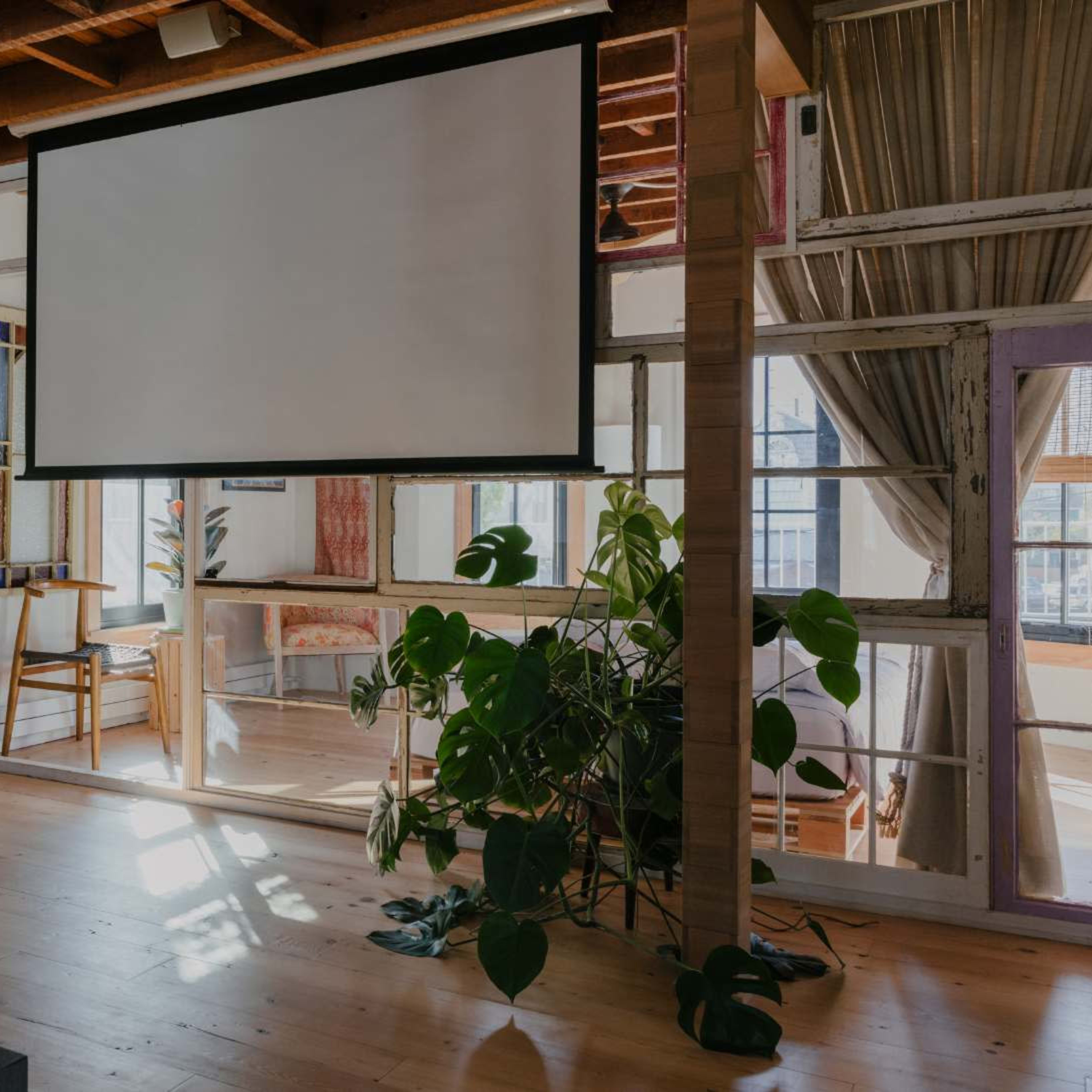 A projector screen hangs from a wooden beam in a sunlit room with wooden floors and large windows, surrounded by greenery and furniture.