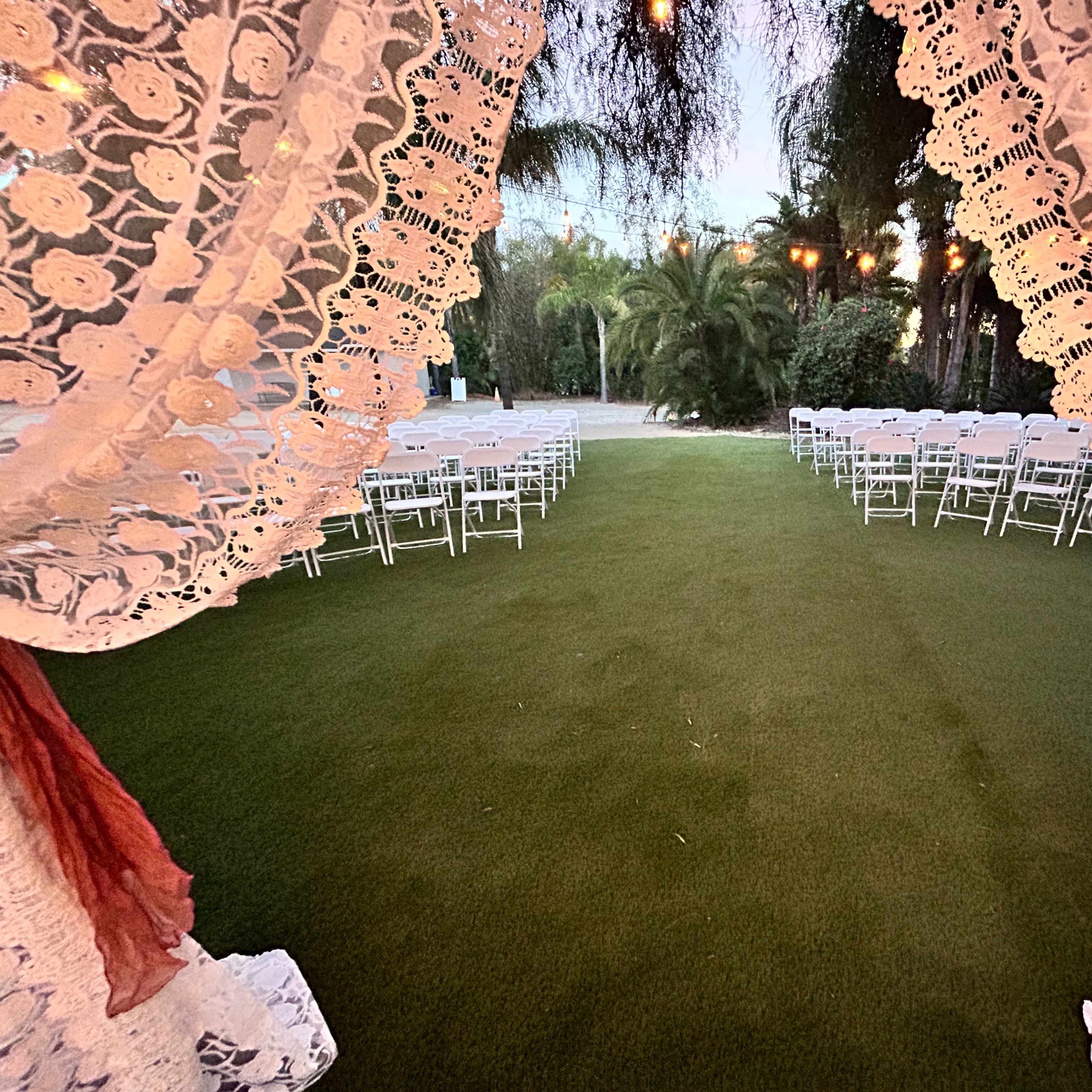 A view through lace curtains framing a grassy aisle leading to rows of white chairs set up for an outdoor event.