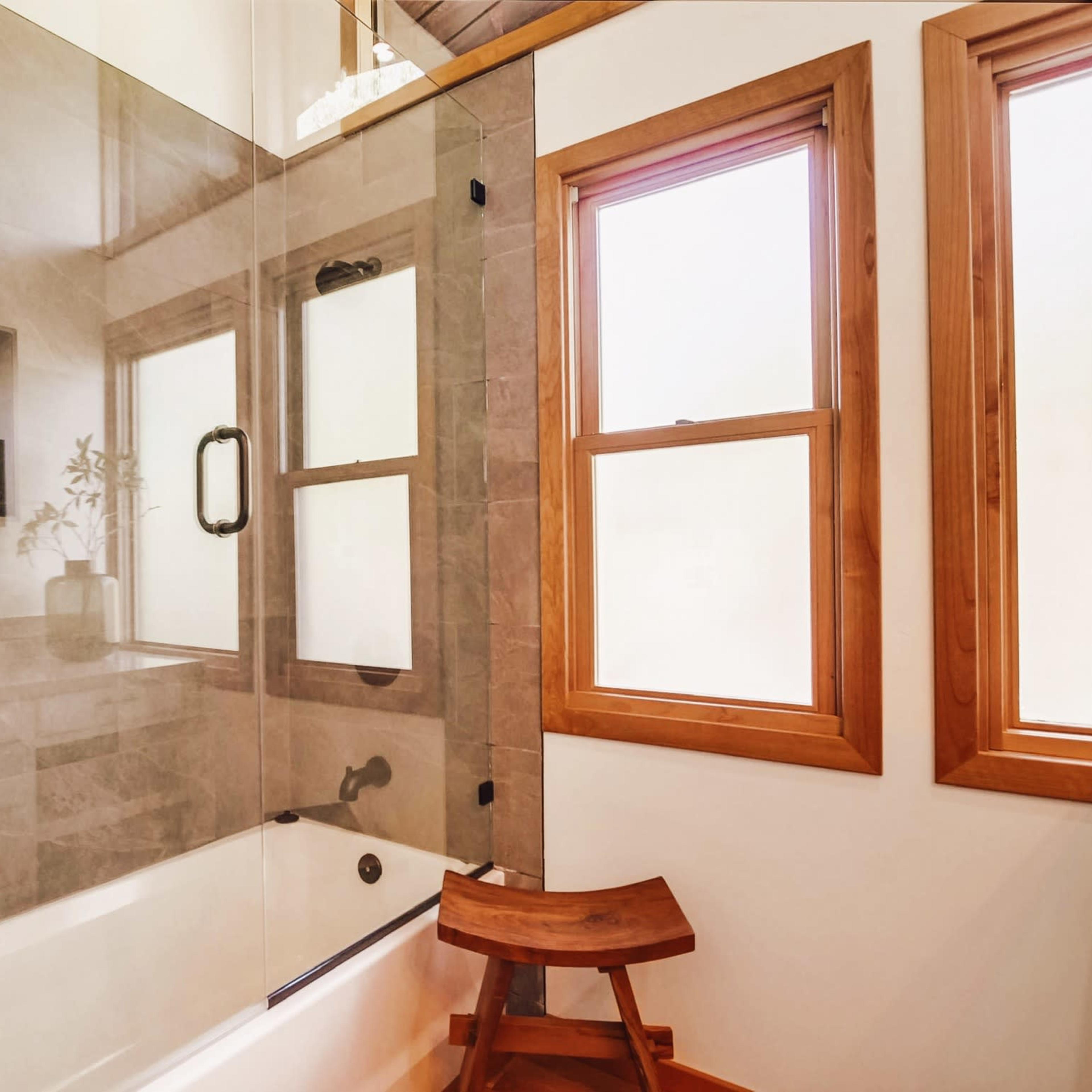 A modern bathroom with a clear glass shower enclosure, wooden windows, and a wooden stool beside a bathtub.