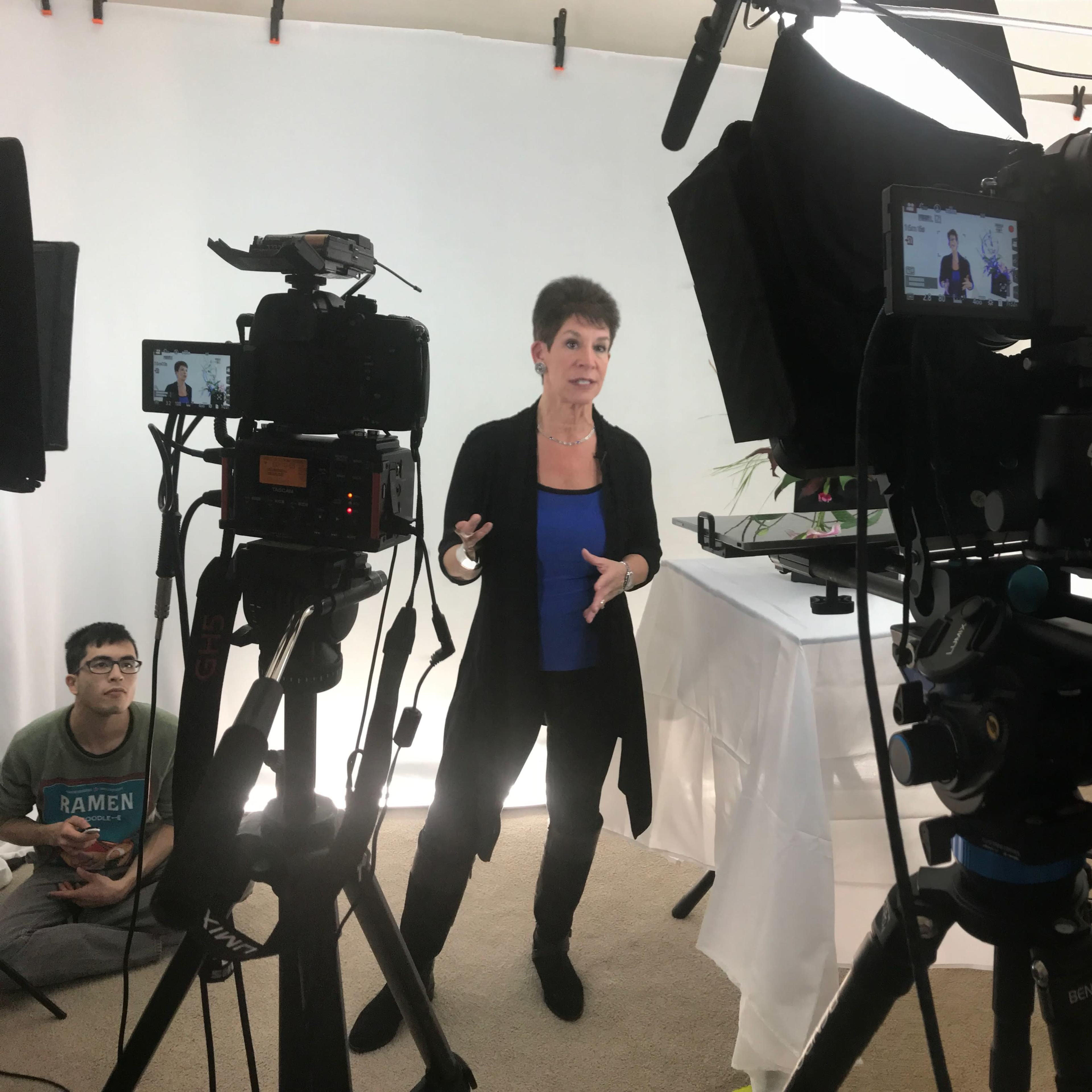 A woman stands in a studio setting, gesturing as she speaks while being recorded by multiple cameras.