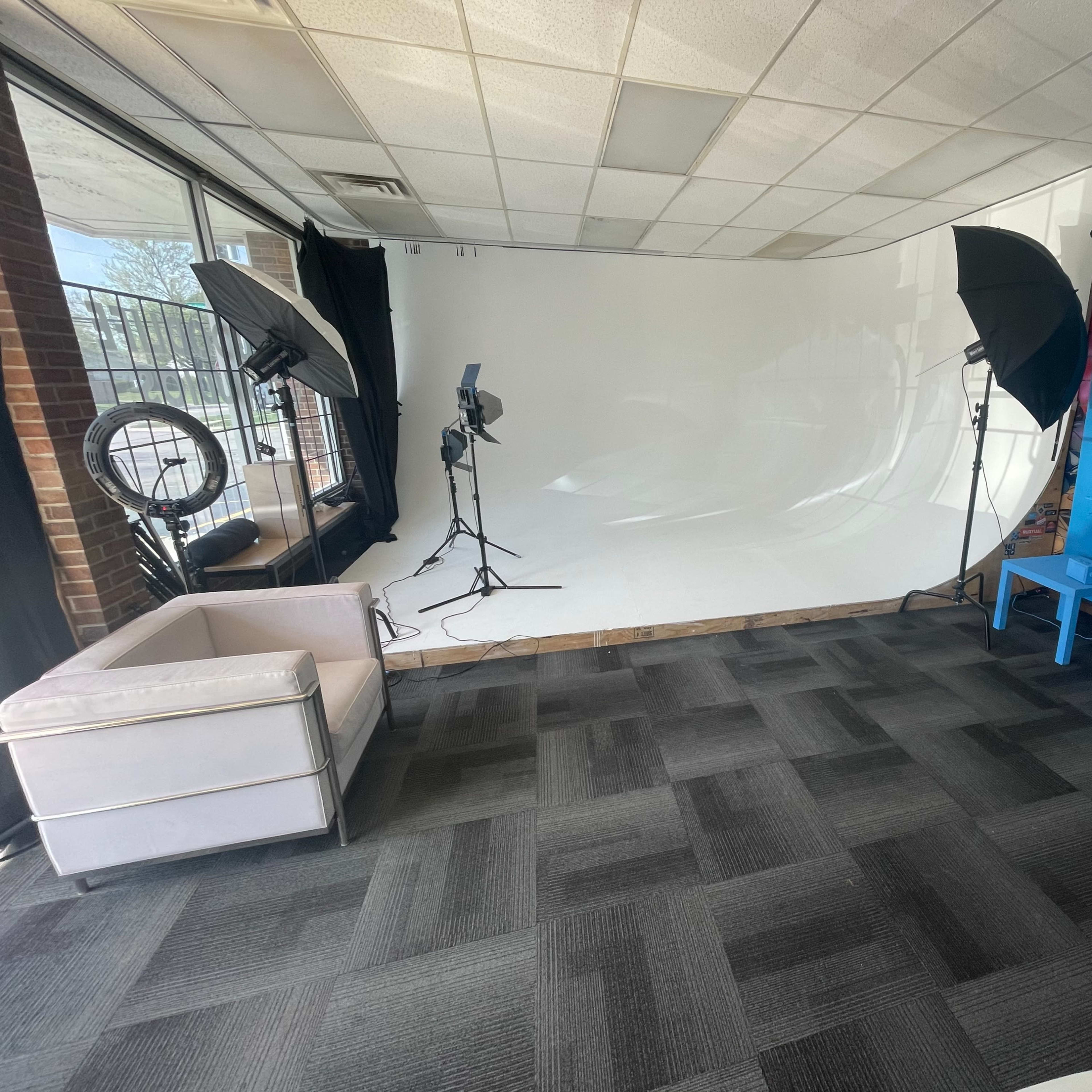 A photography studio with a white backdrop, softbox lights, and a blue wall, featuring a white sofa and two blue chairs.