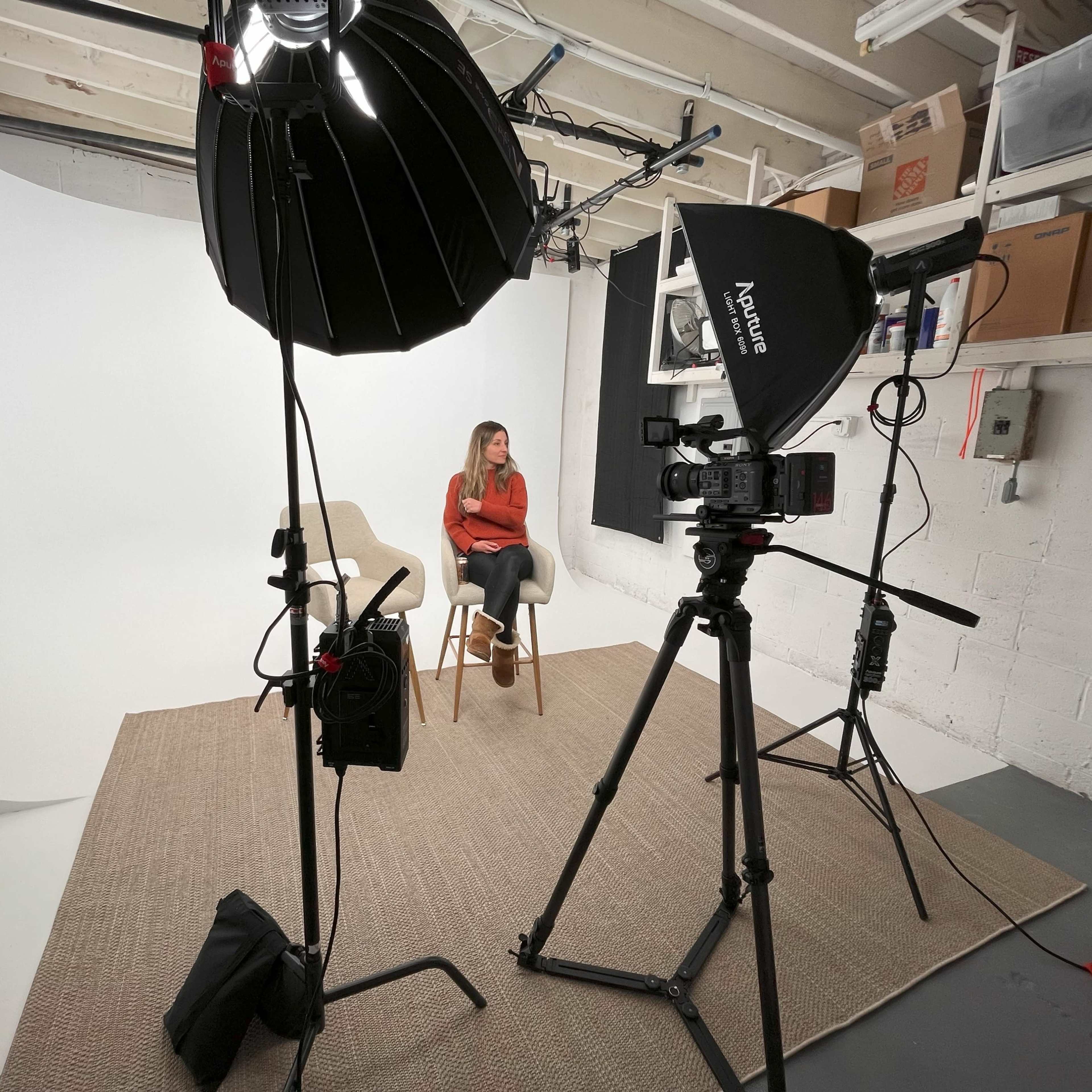 A woman sits on a chair in a photography studio equipped with lighting and a backdrop.