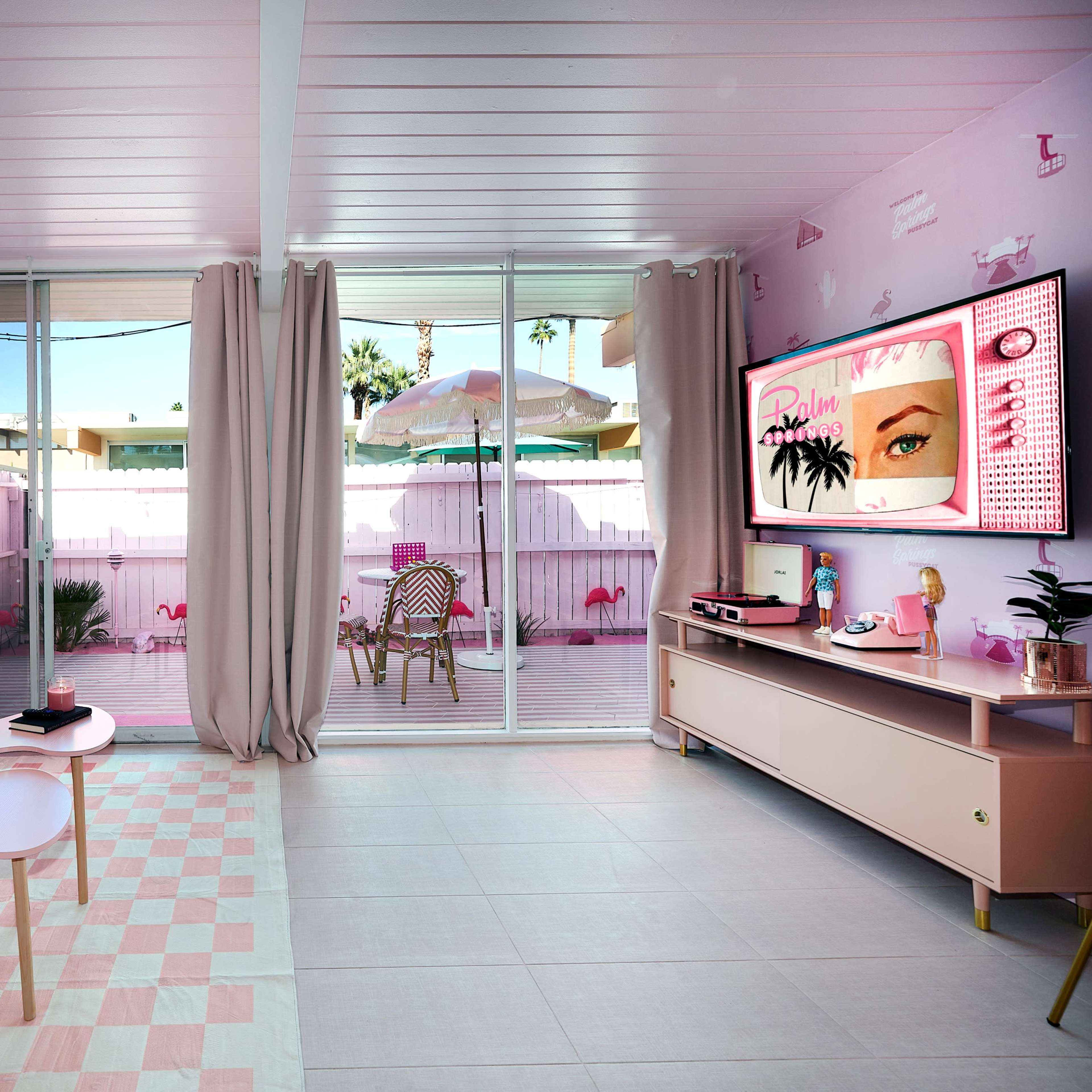 A brightly colored living room features a pink sofa, a small coffee table, and large windows that open to an outdoor patio with flamingo decorations.