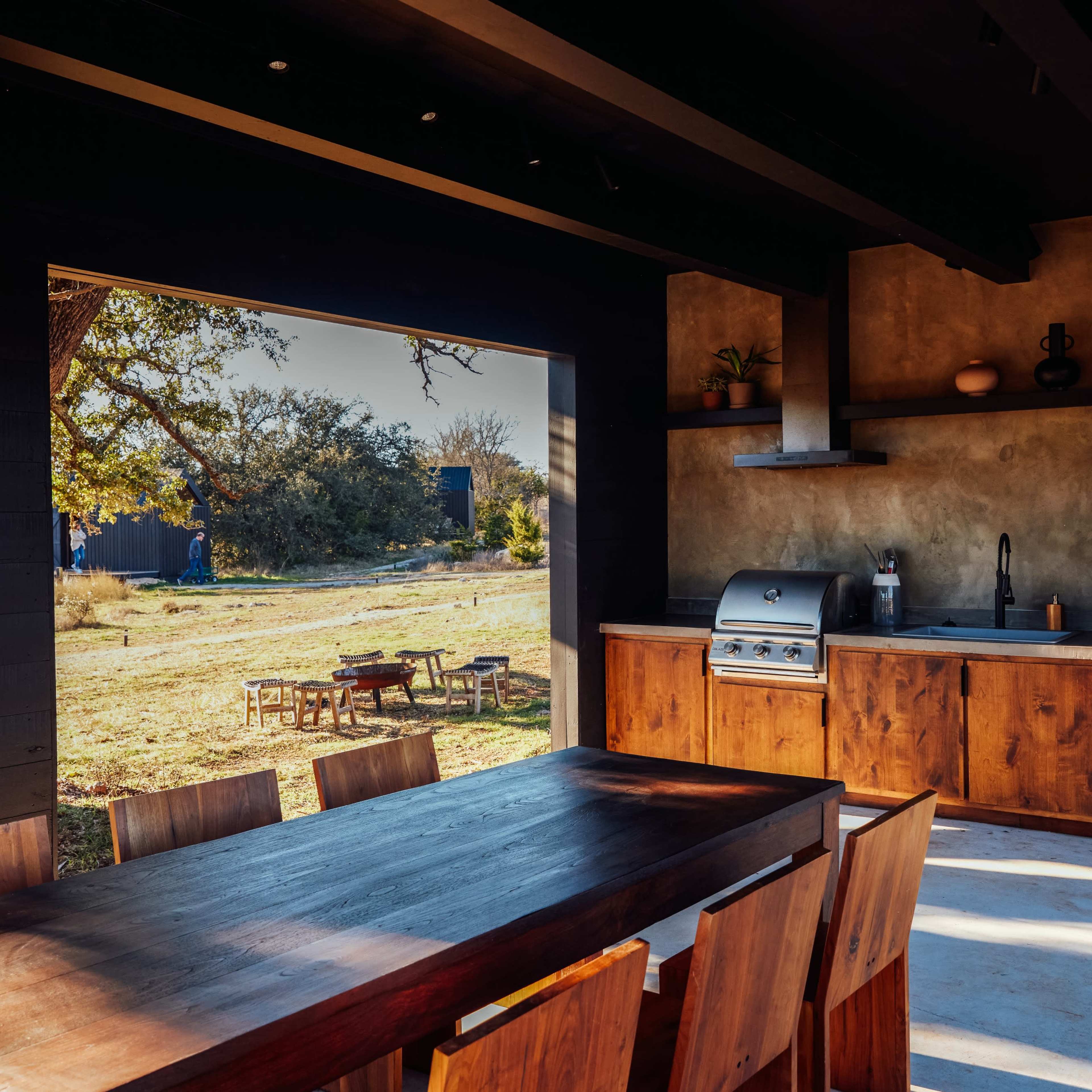 An outdoor kitchen with a wooden dining table and two stainless steel grills, set against a backdrop of a grassy area and trees.