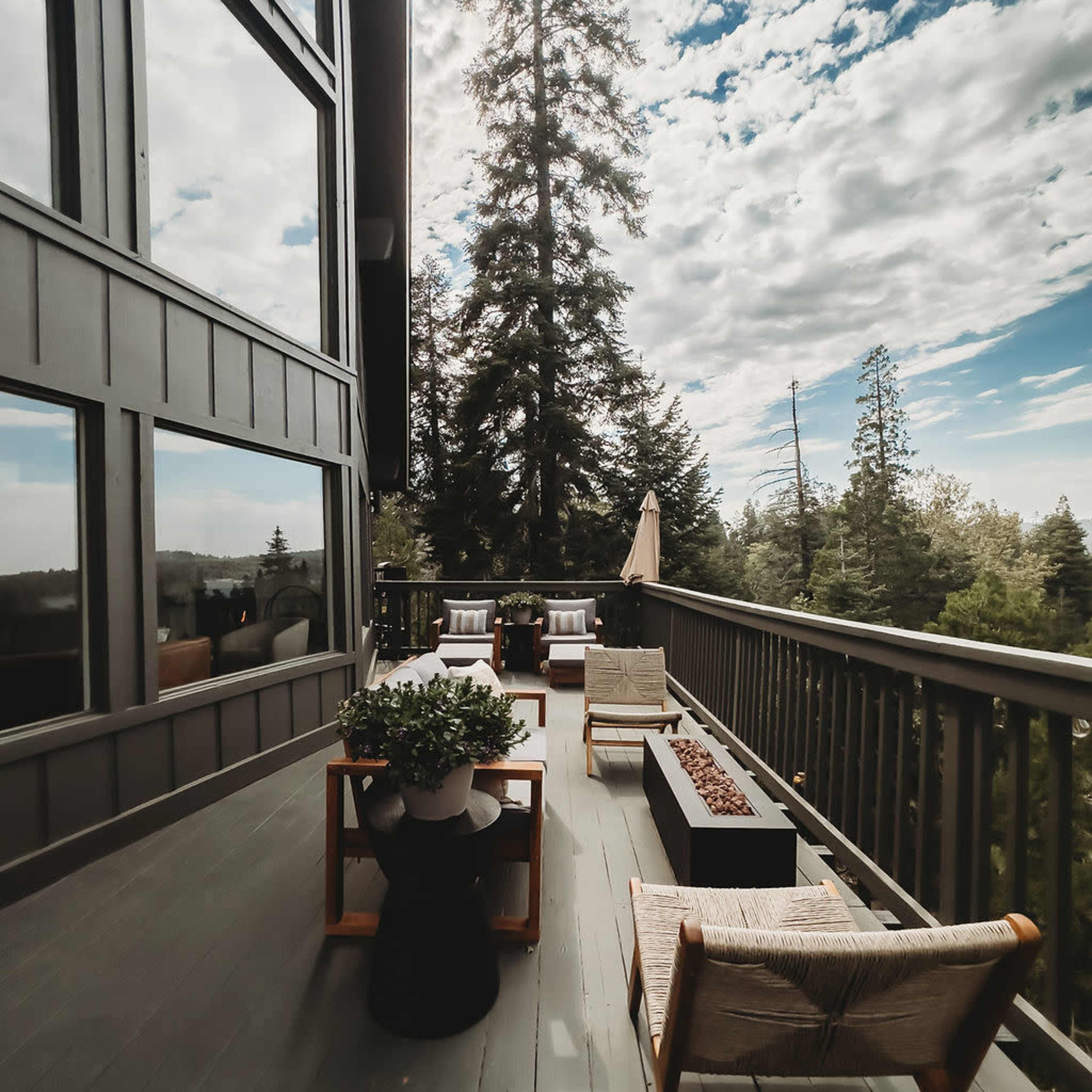 A spacious balcony features wooden furniture, potted plants, and a fire pit, surrounded by tall trees and a cloudy sky.