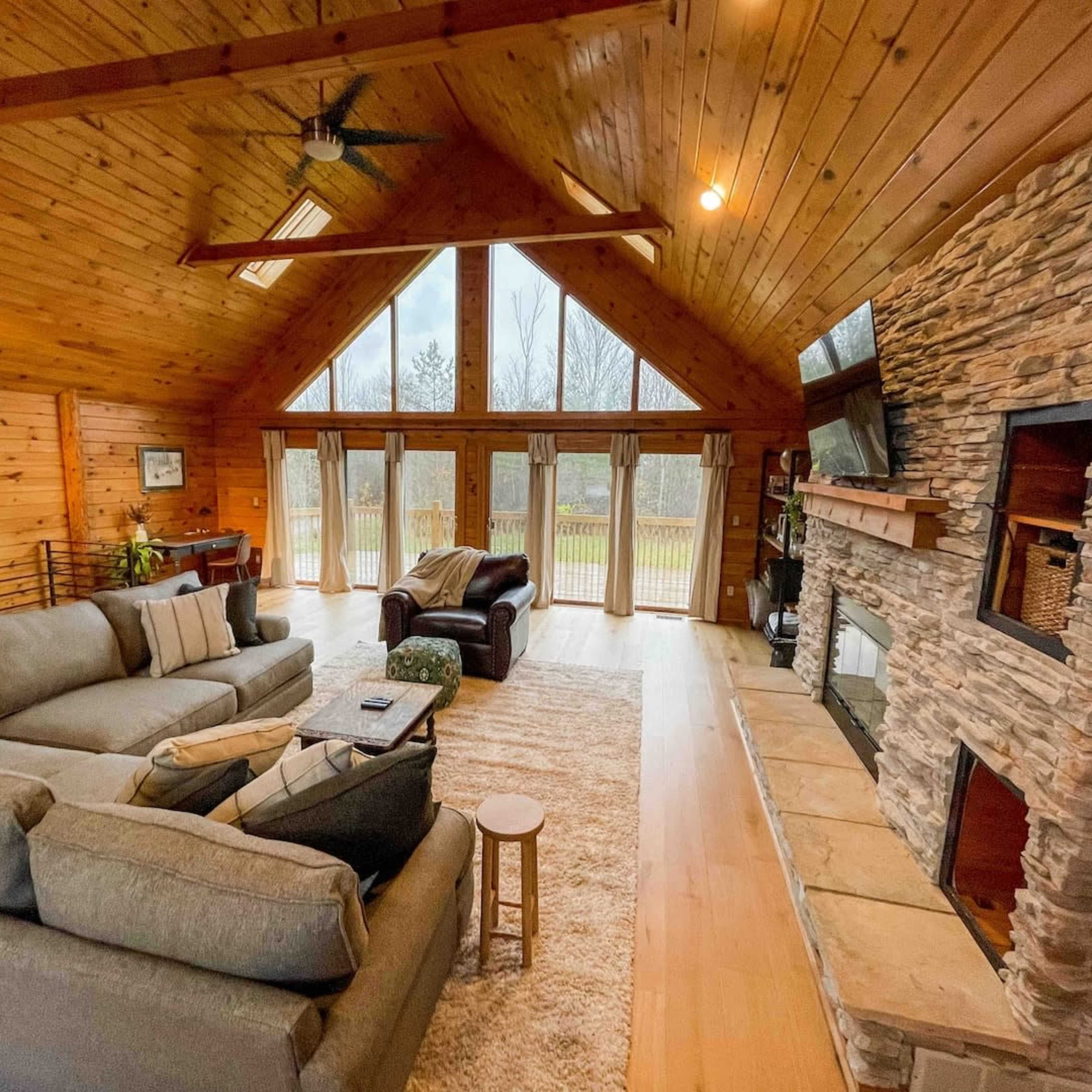 The living room features a large sectional sofa, a stone fireplace, and large windows framed by wooden walls, allowing natural light to illuminate the space.