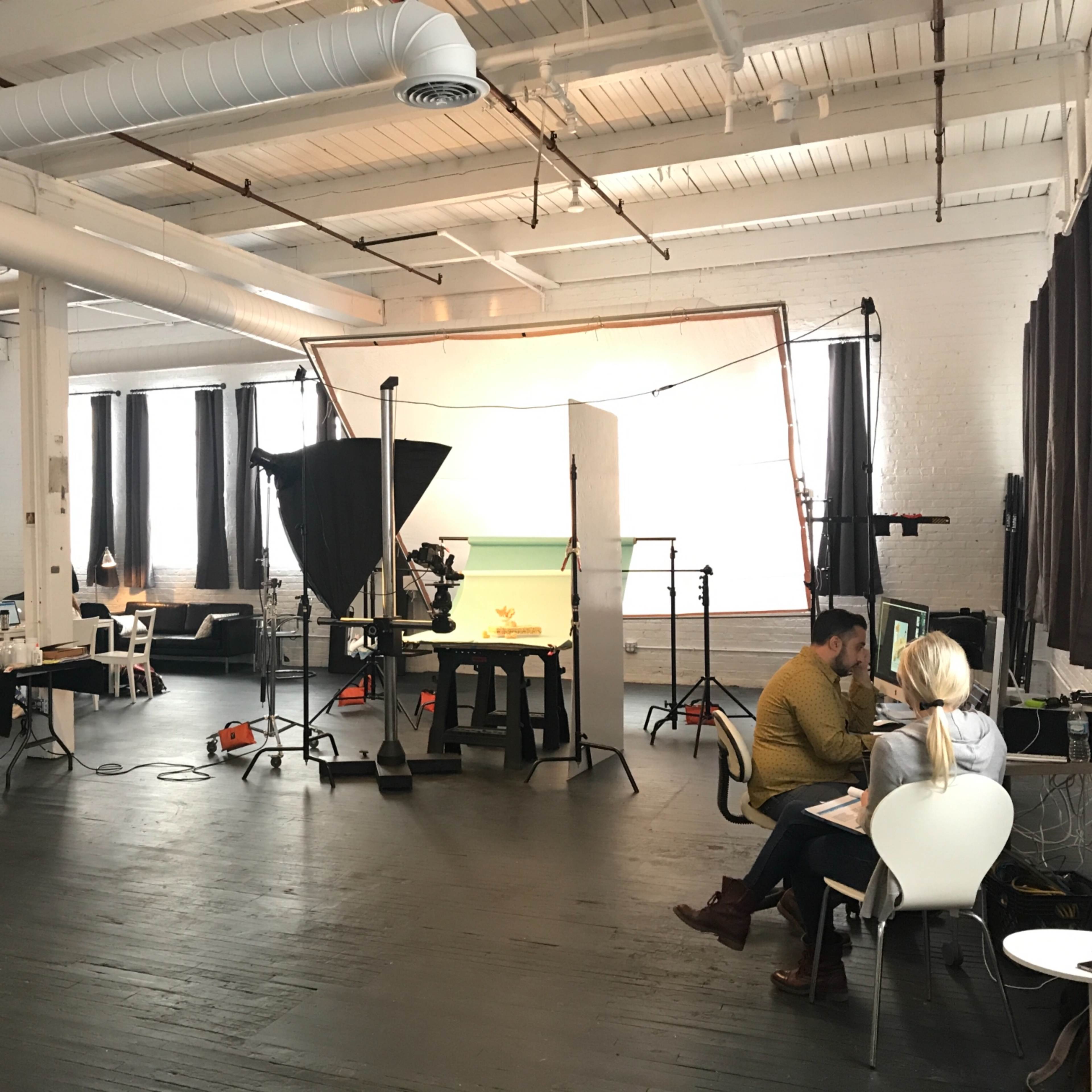 A spacious studio with various workstations, lighting equipment, and two people seated at a table engaged in conversation.