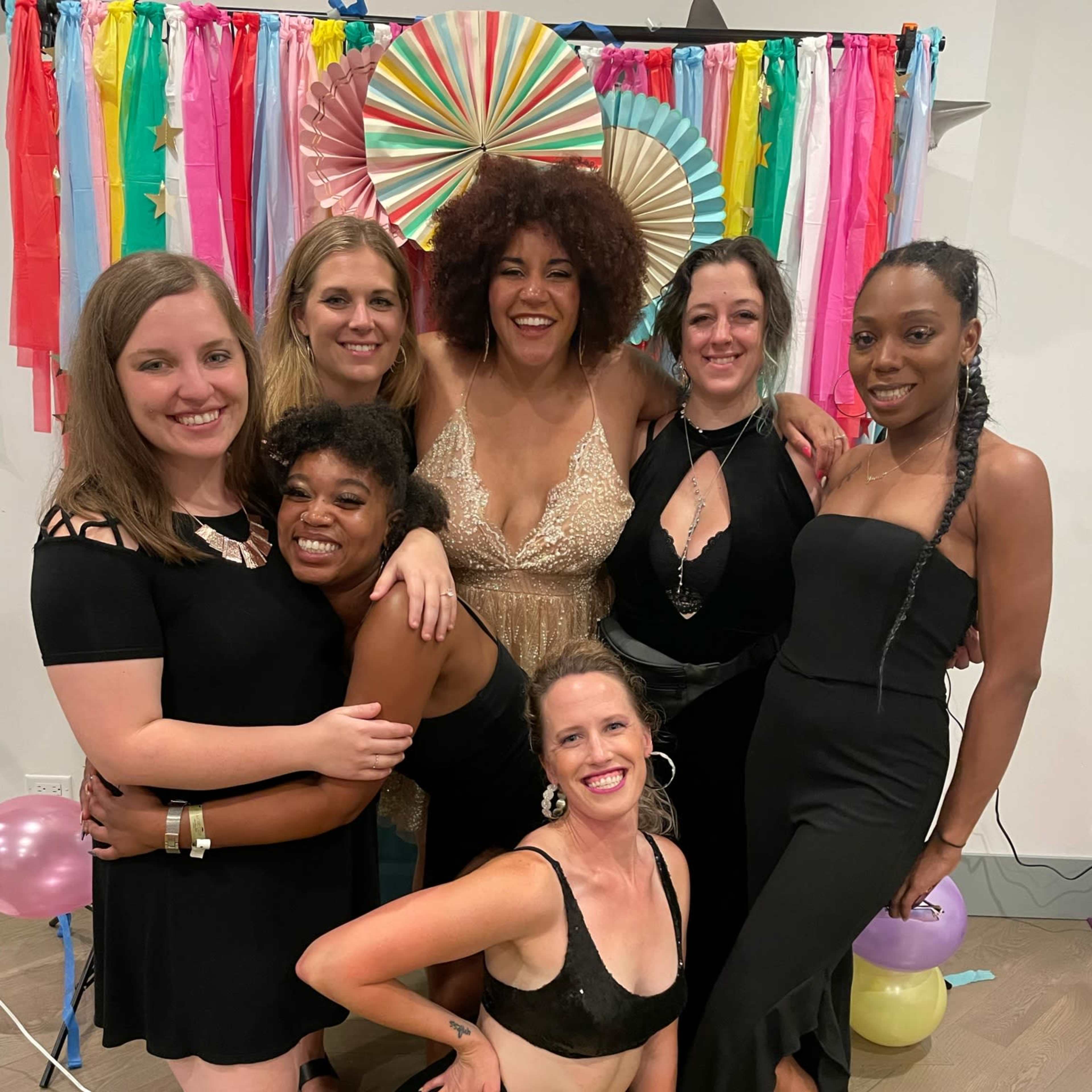 A group of six women poses together, wearing black dresses and celebrating in front of a colorful backdrop.
