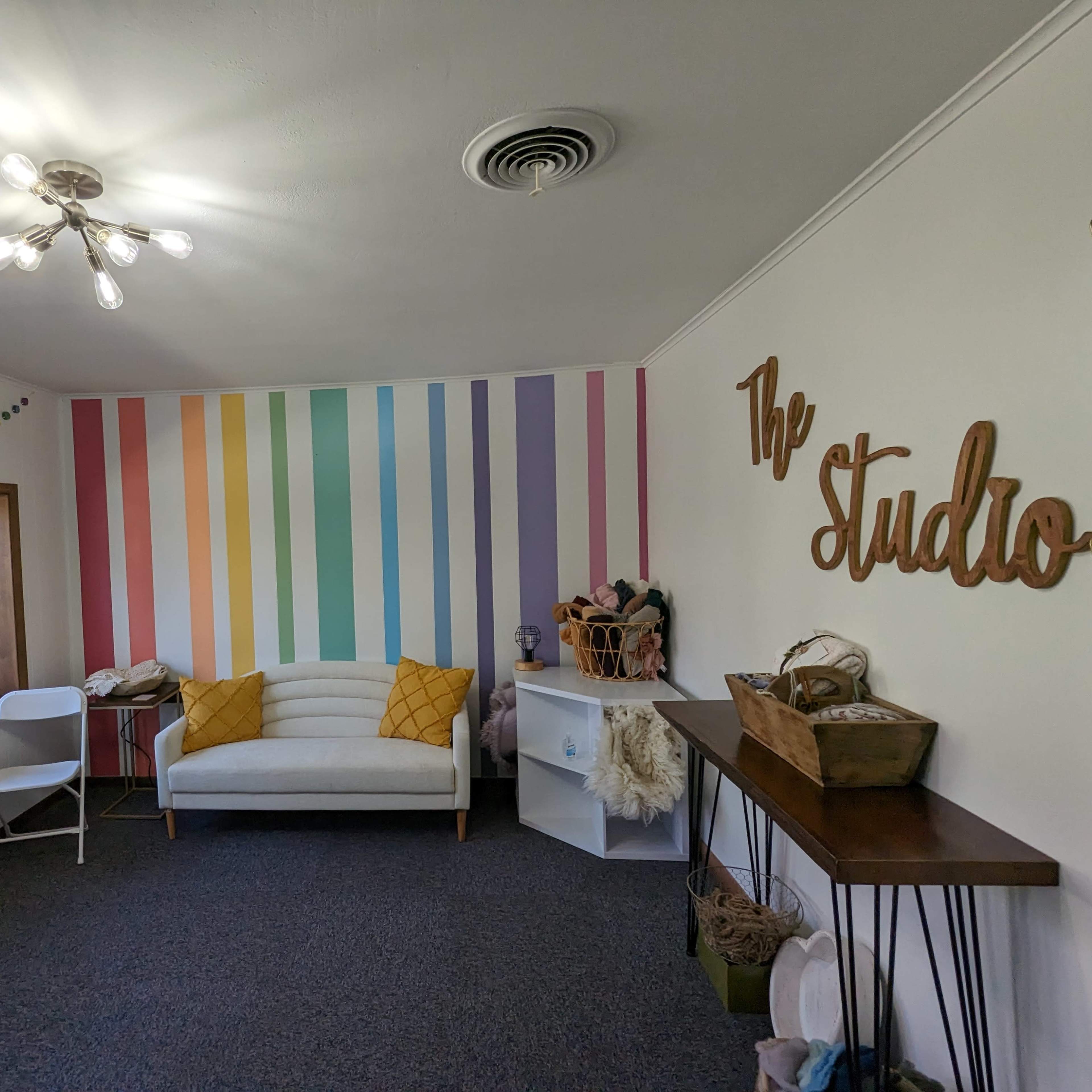 A bright room features a white couch with yellow cushions, colorful striped walls, and a decorative wooden sign that reads "The Studio."