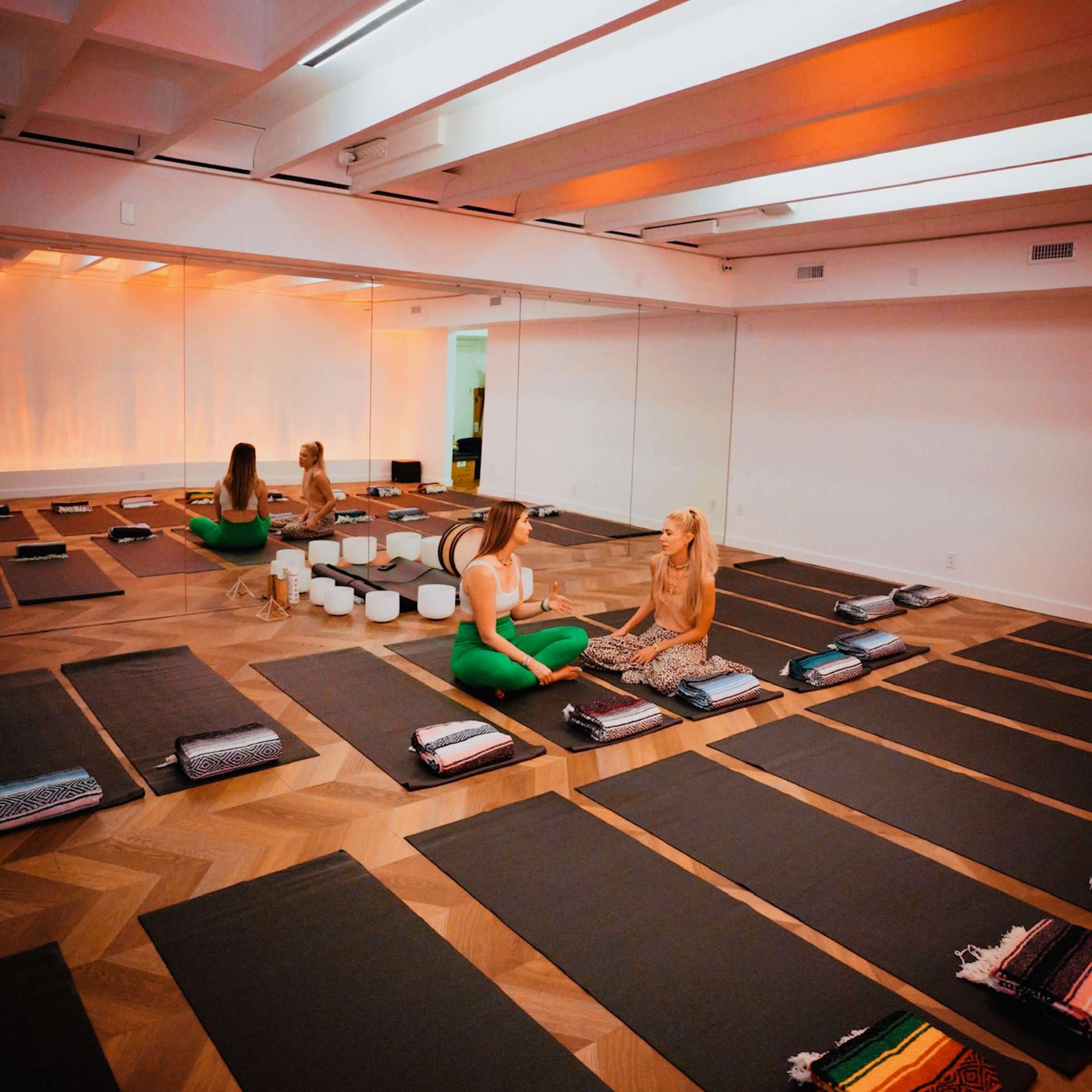 A yoga studio features several exercise mats arranged on a wooden floor, with two women sitting and conversing in a well-lit space.