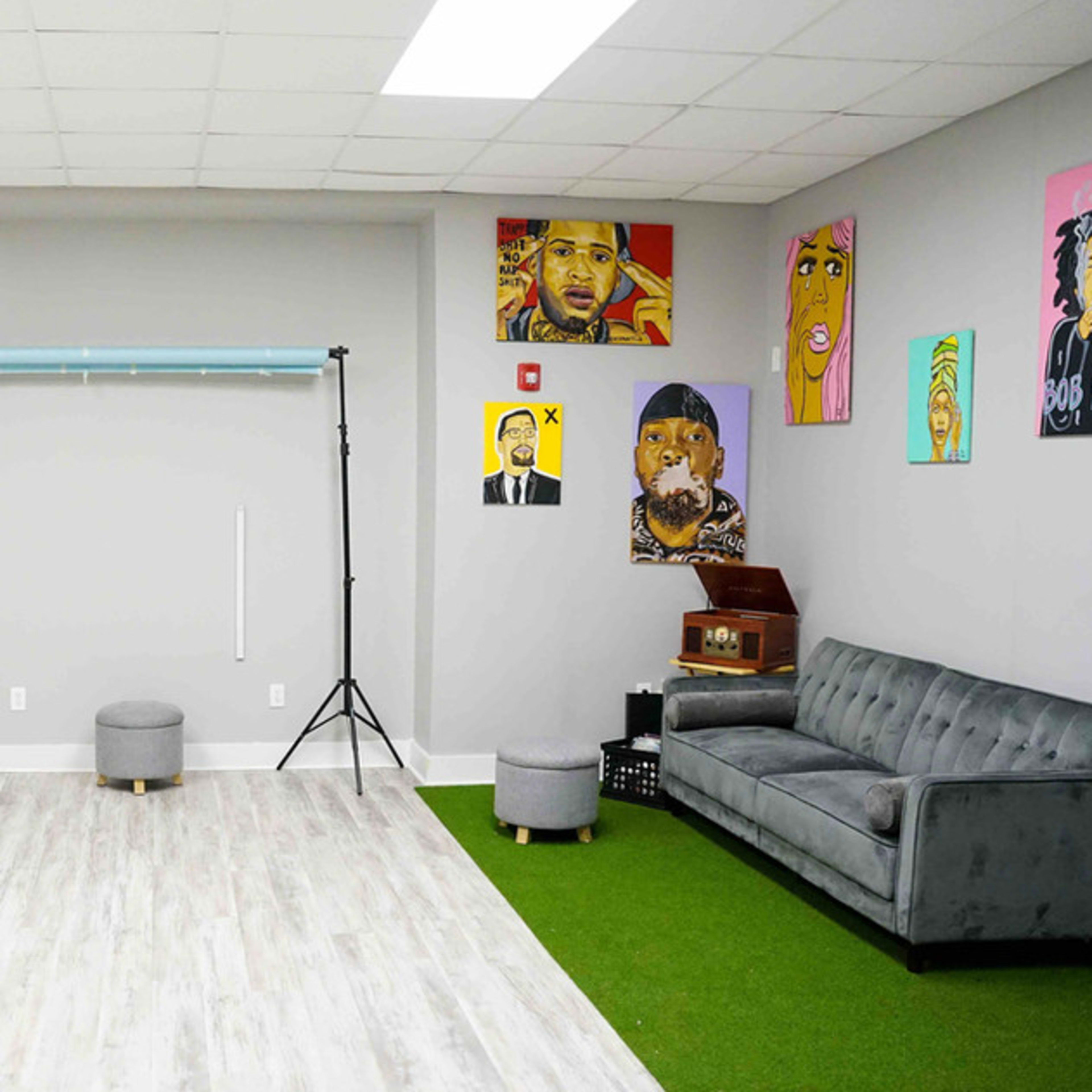 A spacious studio with colorful backdrop rolls, a photography setup, artwork on the walls, a vinyl record player, and a gray couch on a green area rug.