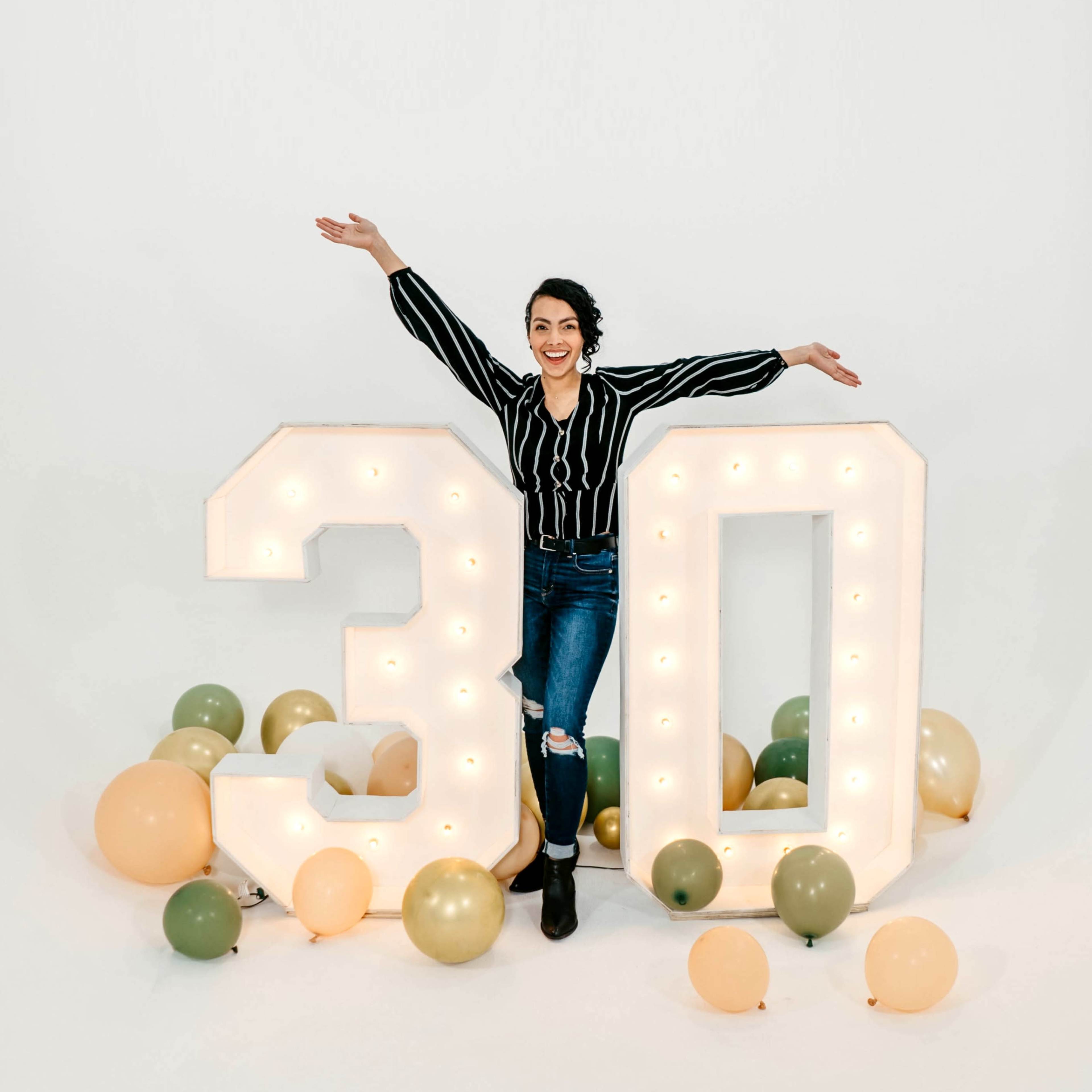 A woman stands beside large illuminated "30" numbers, surrounded by colorful balloons.