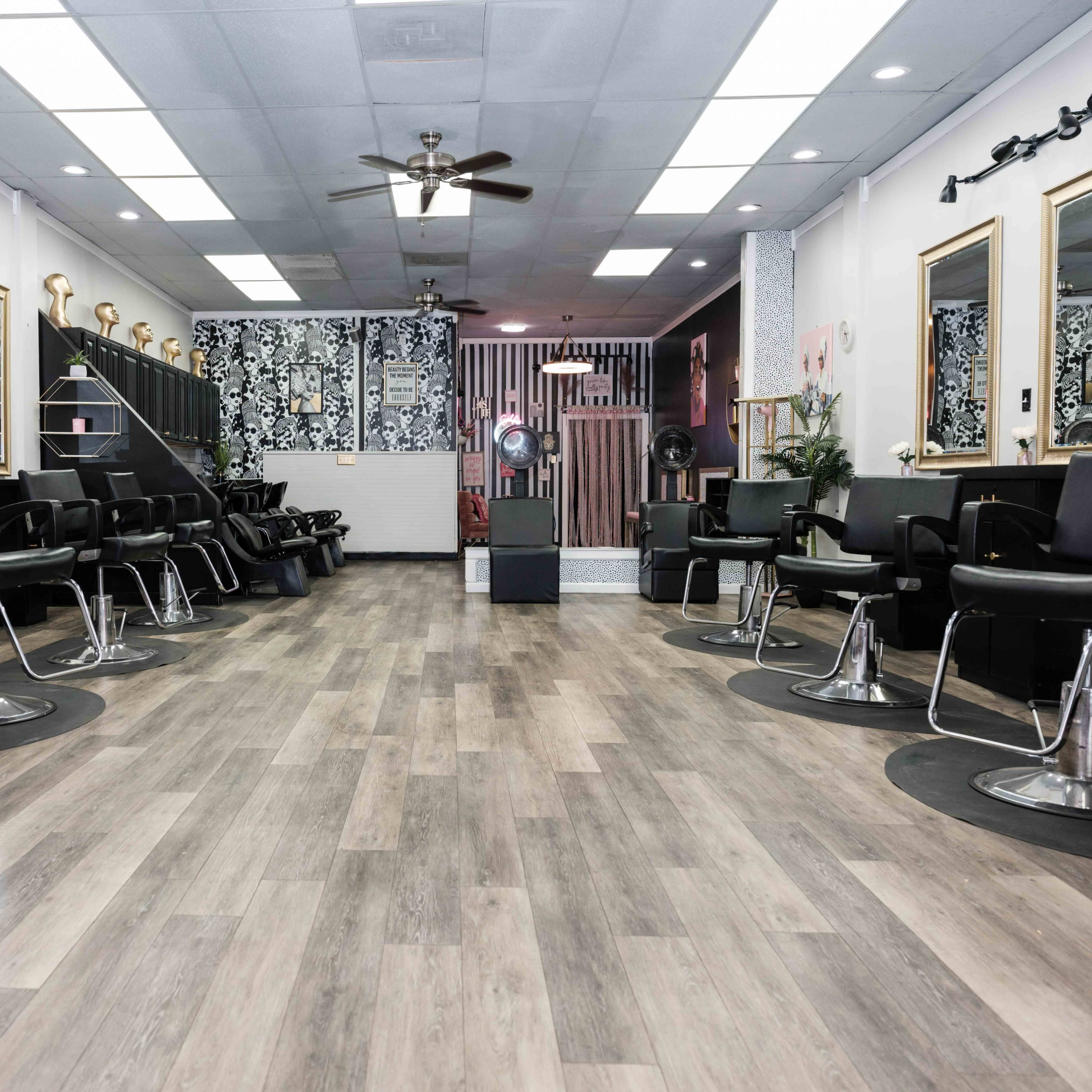 A modern hair salon featuring black styling chairs, large mirrors, and a spacious wooden floor.