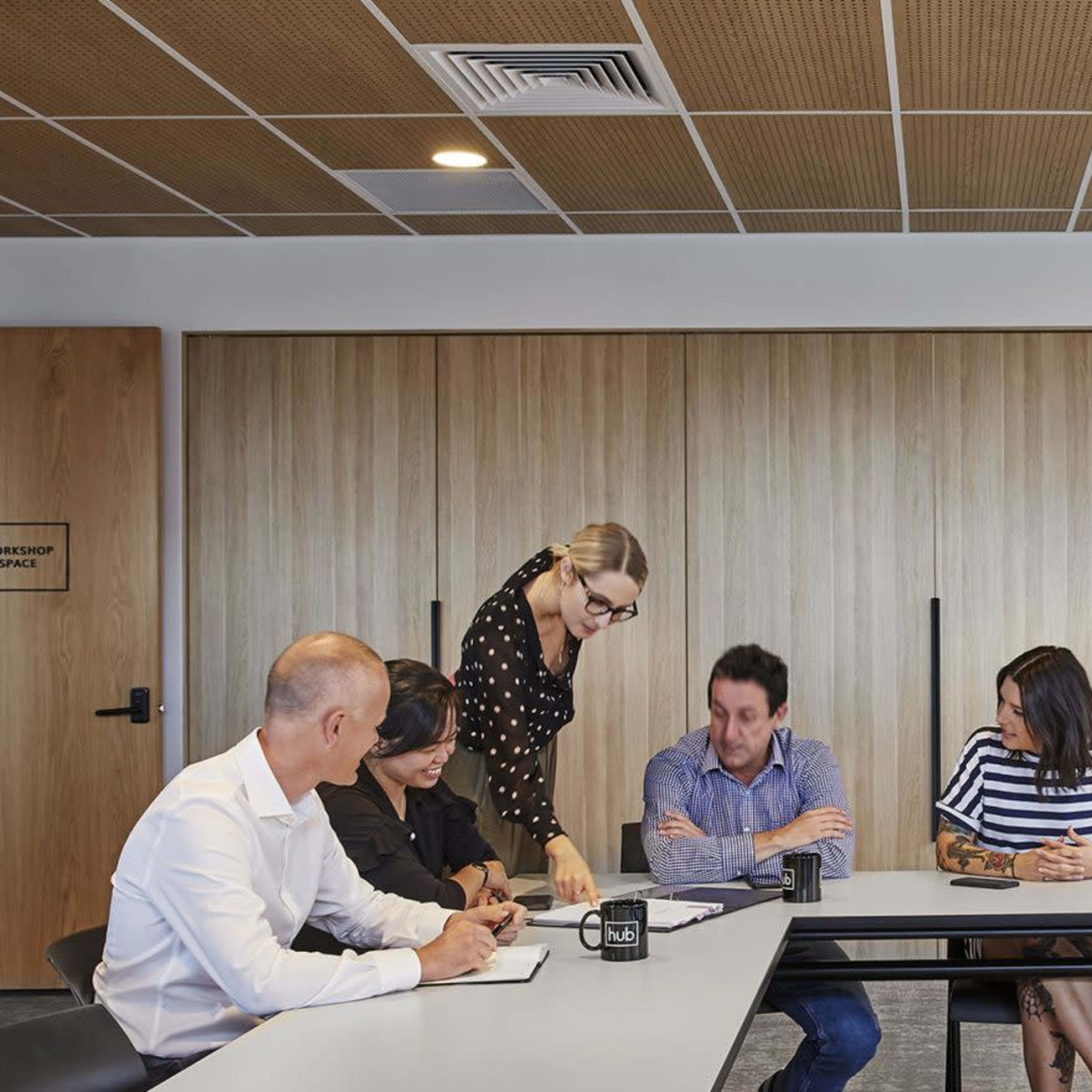 A group of professionals is seated around a long table in a meeting room, while one person stands and addresses the group.