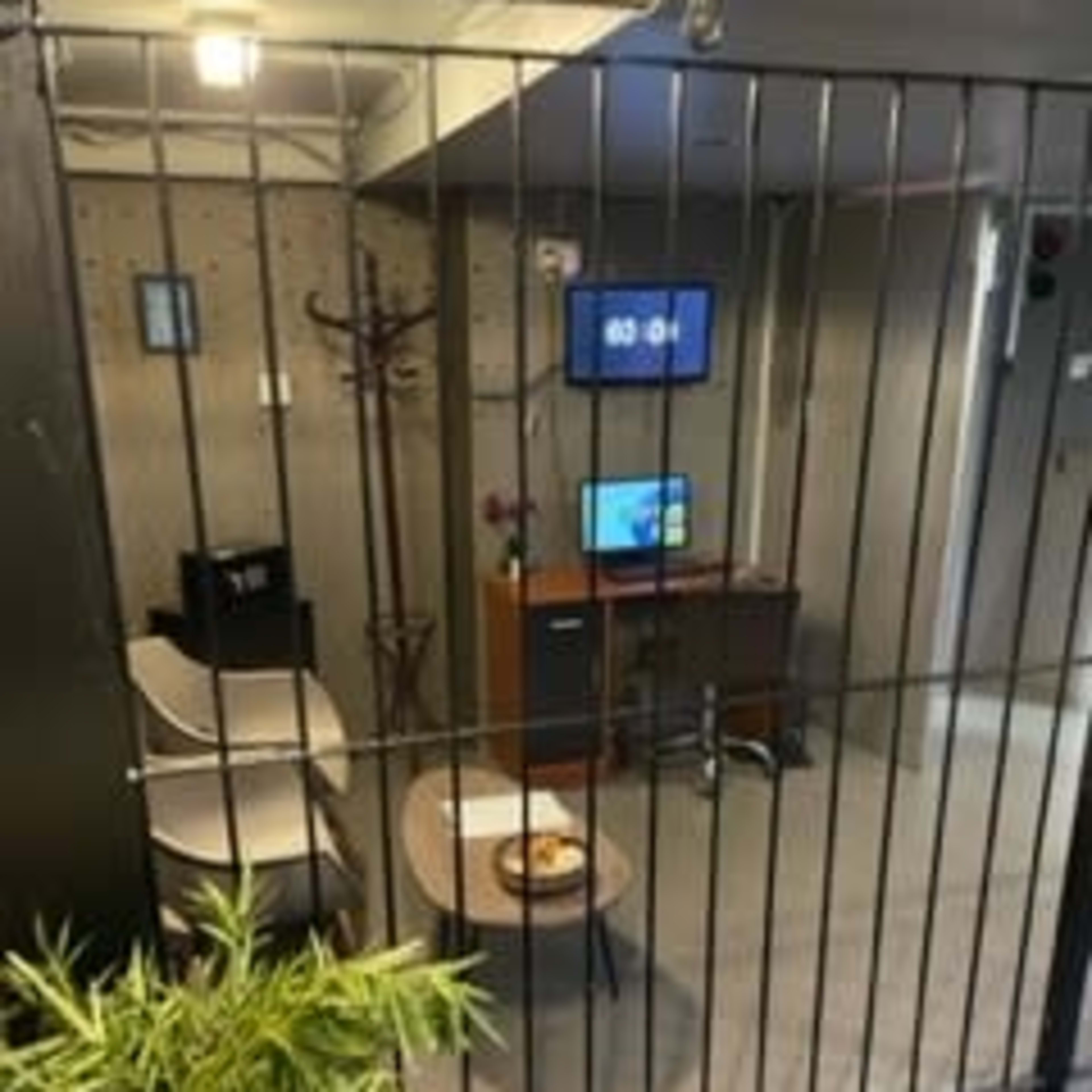 A small, enclosed office space with a desk, two chairs, a television, and a potted plant, all behind a metal grid barrier.
