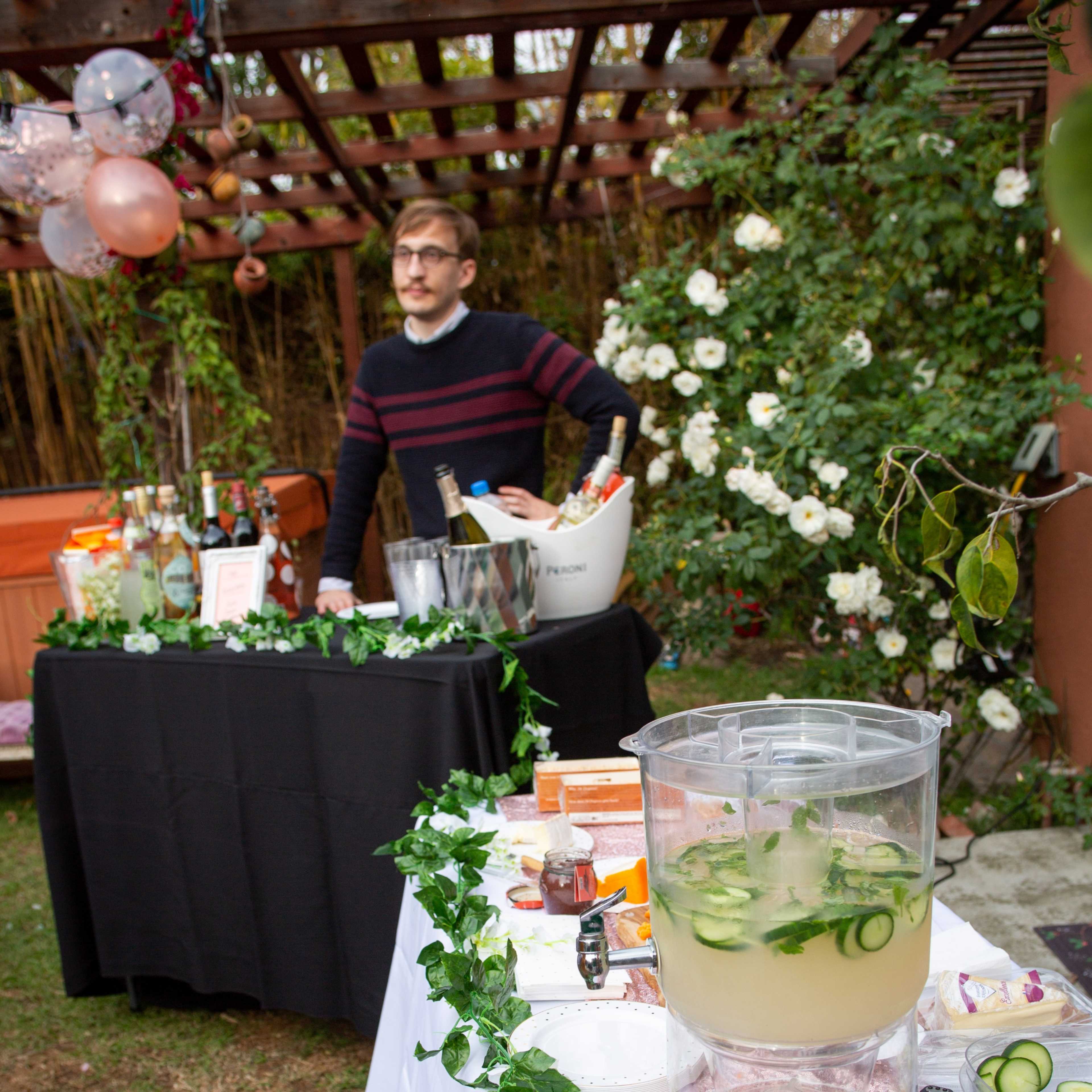 A man stands behind a bar setup featuring drinks and snacks in a garden adorned with flowers and decorations.
