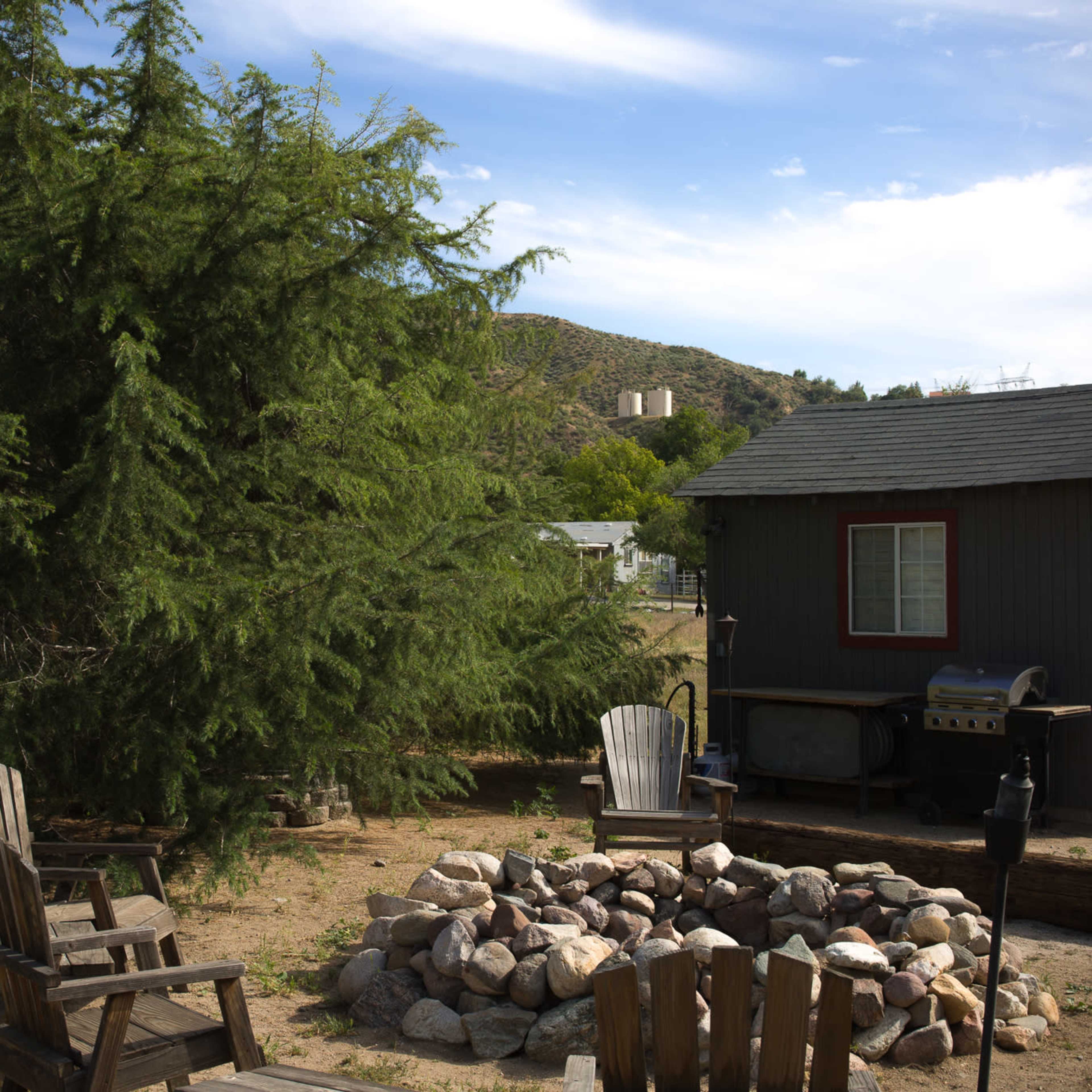 A rustic outdoor setting with a stone fire pit surrounded by wooden chairs, near a small gray house and large trees, against a hilly background.