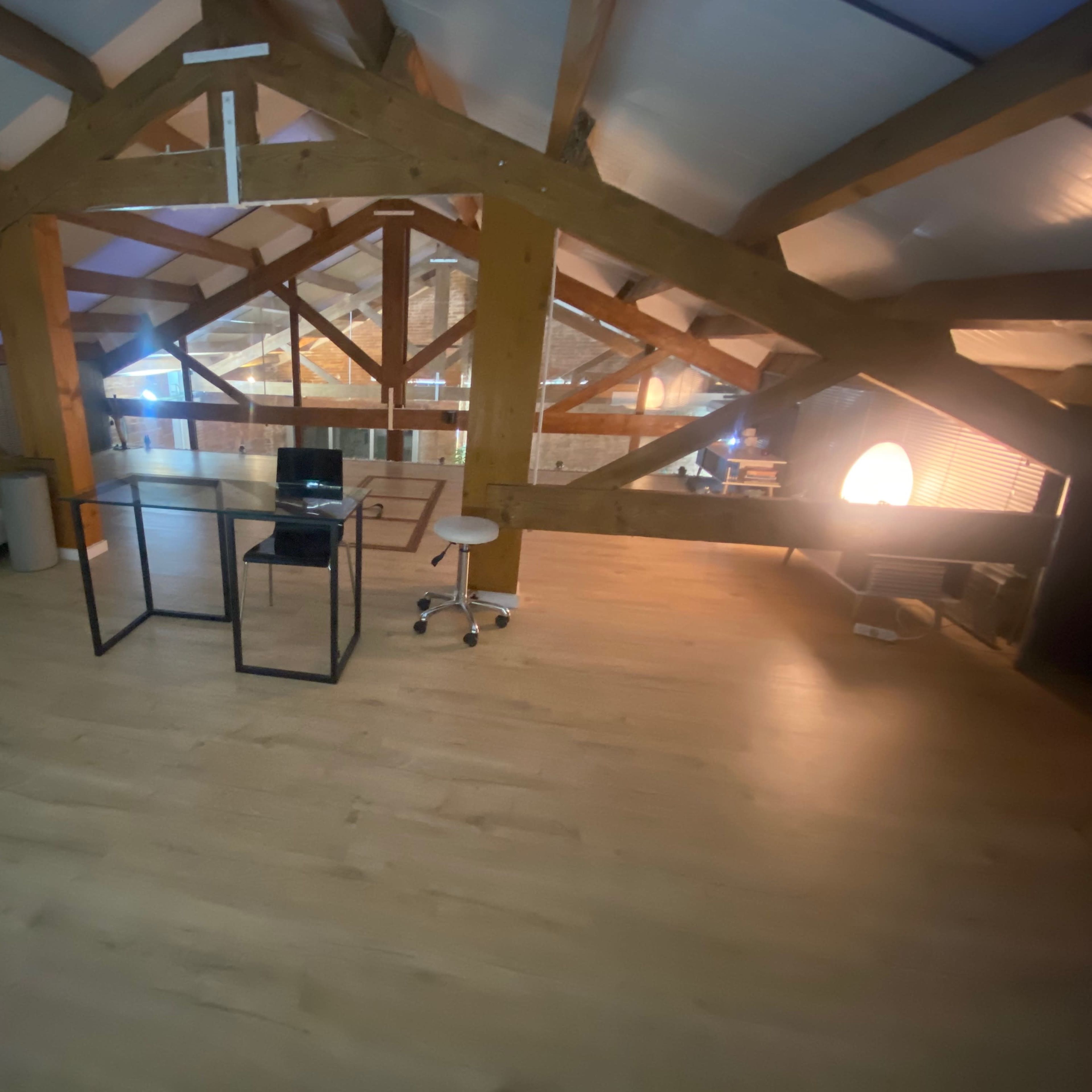 A spacious loft with wooden beams, a pale wooden floor, a computer setup on a glass table, and a few chairs.