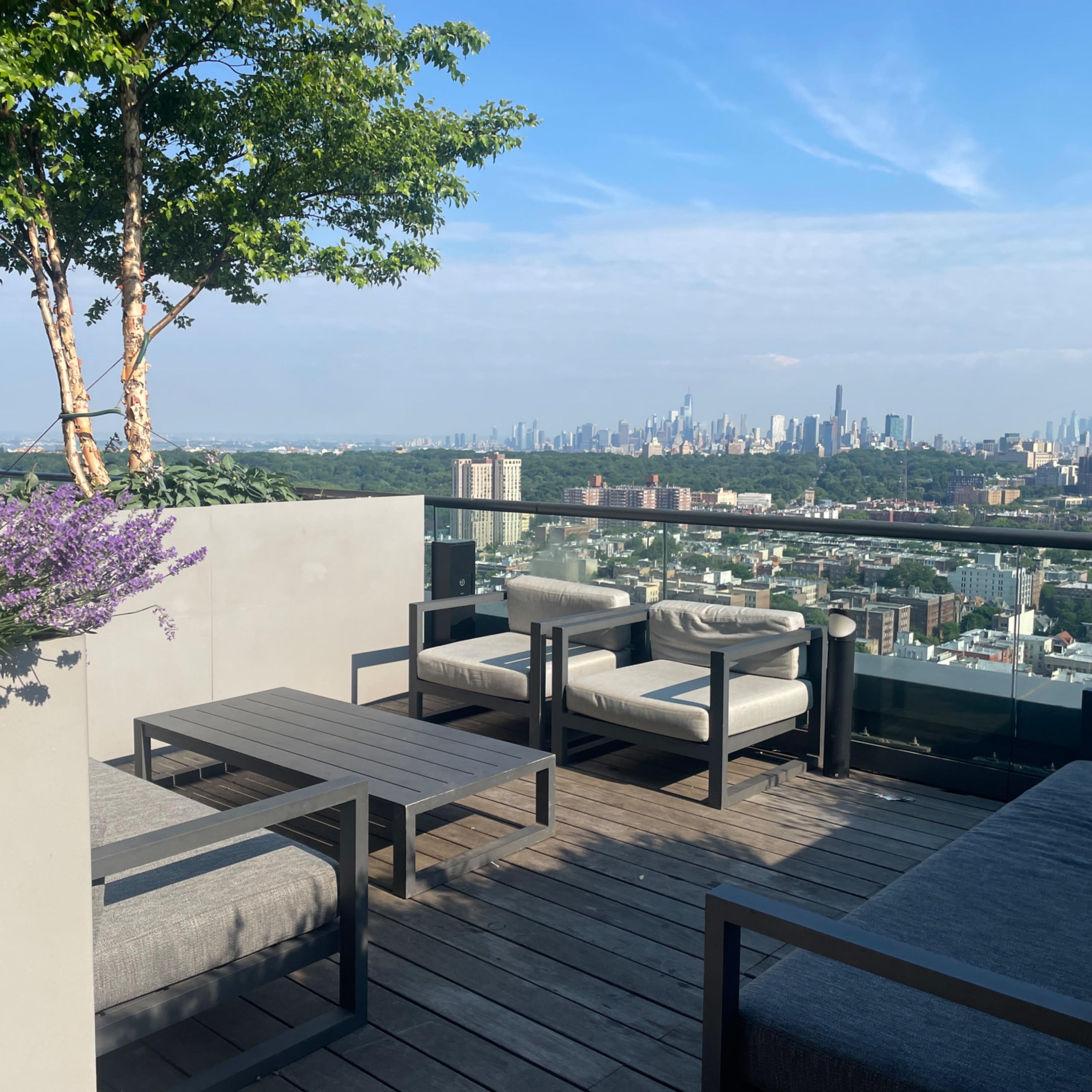 A rooftop patio features modern seating and a view of a city skyline against a clear blue sky.