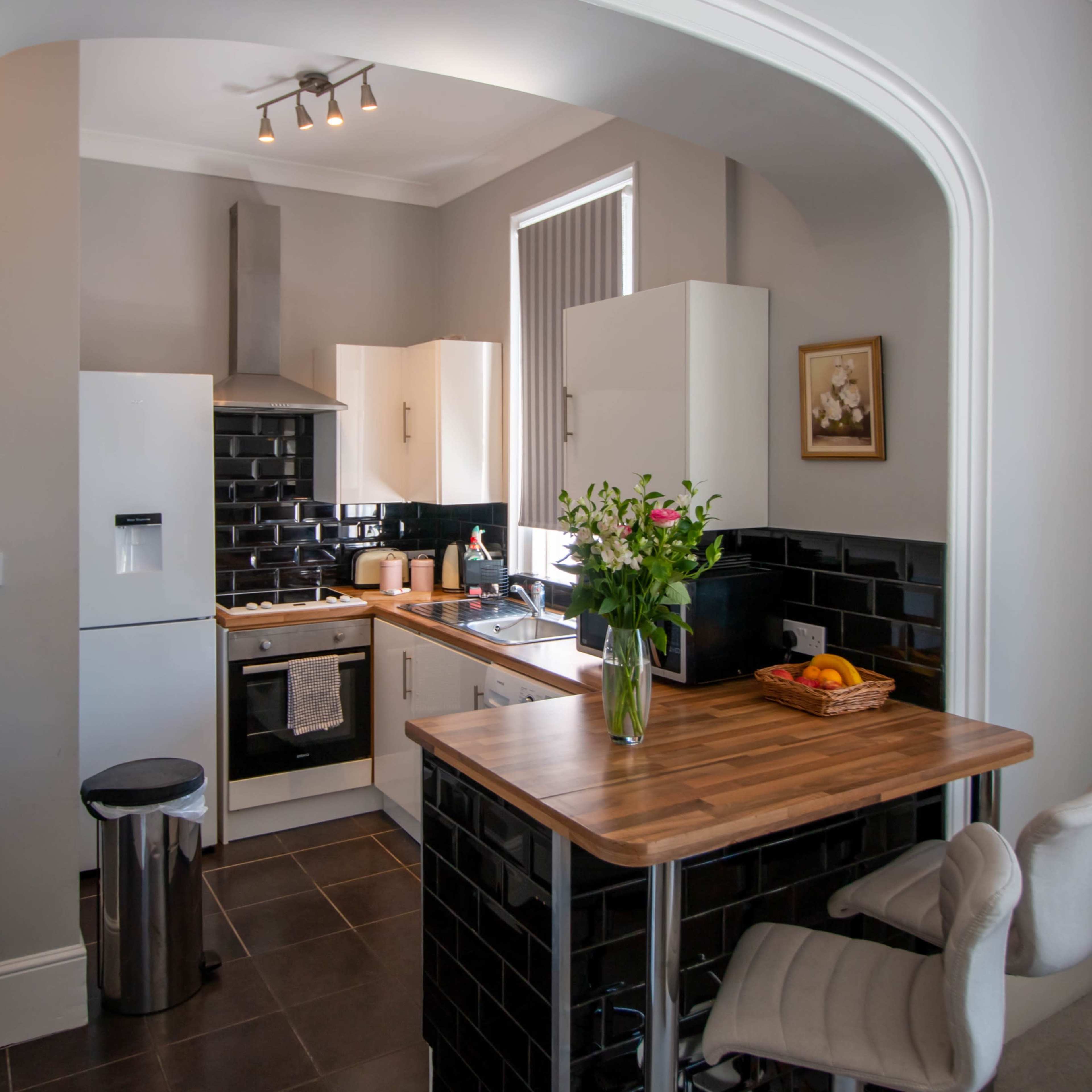A modern kitchen features white cabinets, a black tiled backsplash, and a wooden table with a vase of flowers and a bowl of fruit.