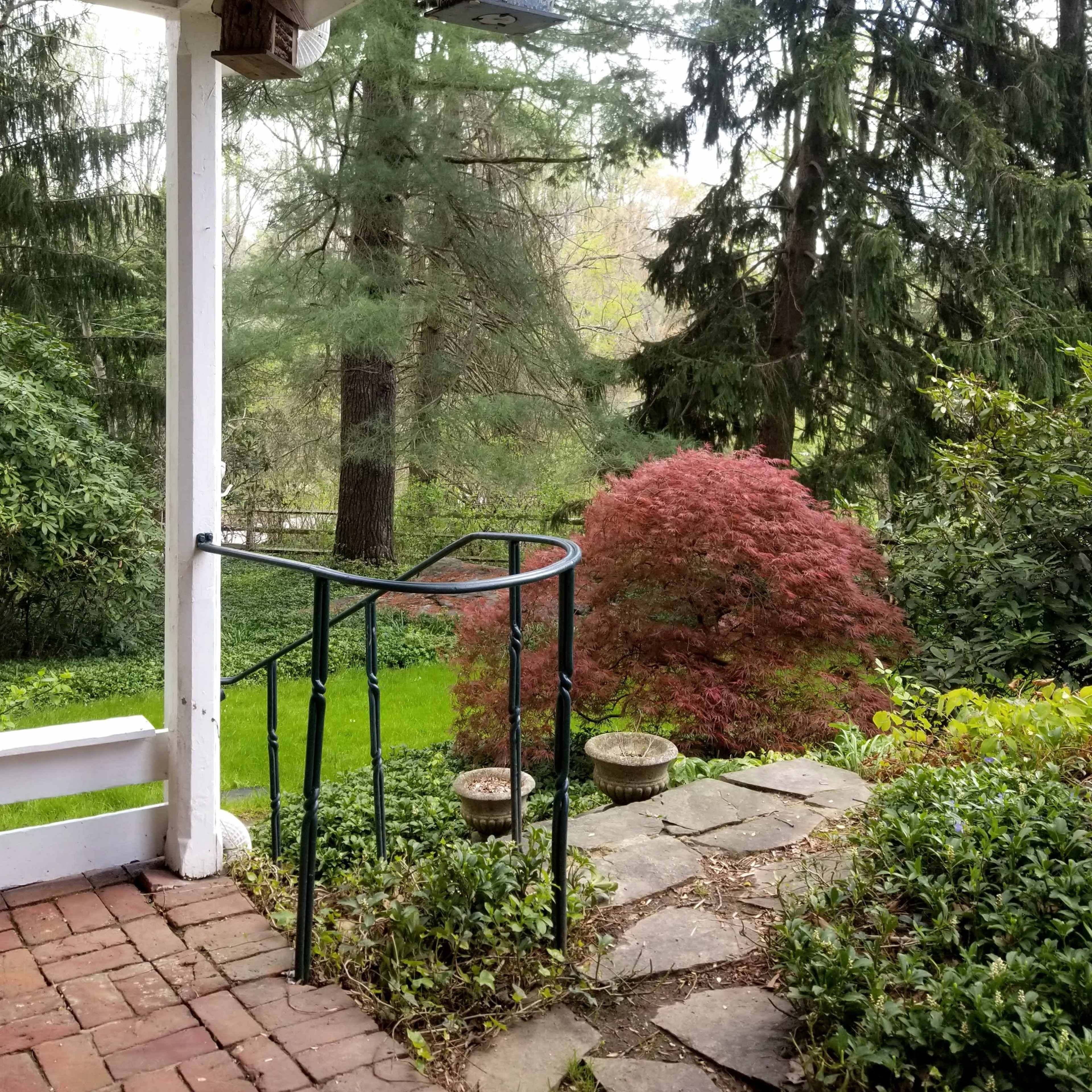 A view from a covered porch, featuring a curved metal railing, a stone pathway, and various trees and shrubs in the garden.