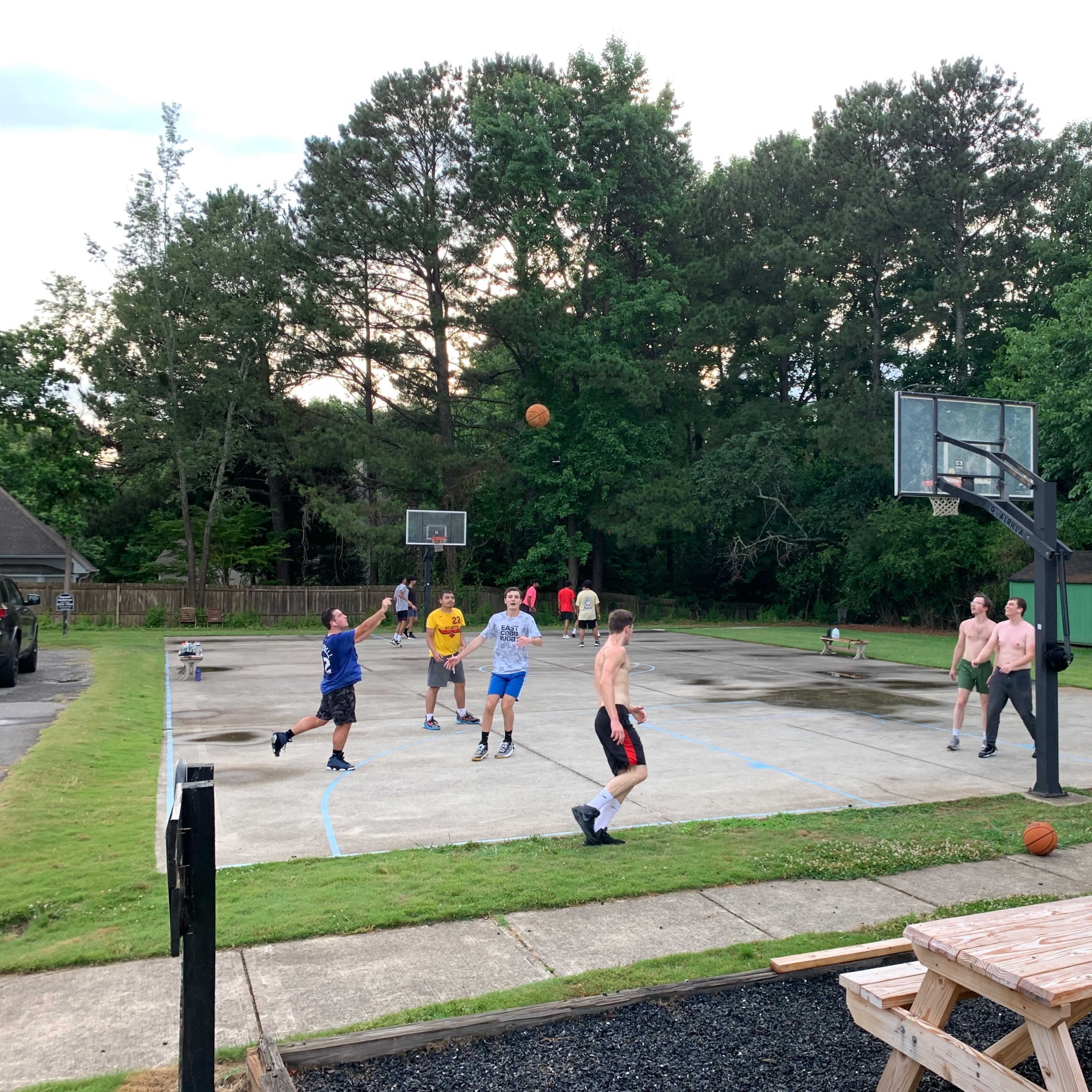 A group of people plays basketball on an outdoor court surrounded by trees and a picnic table nearby.