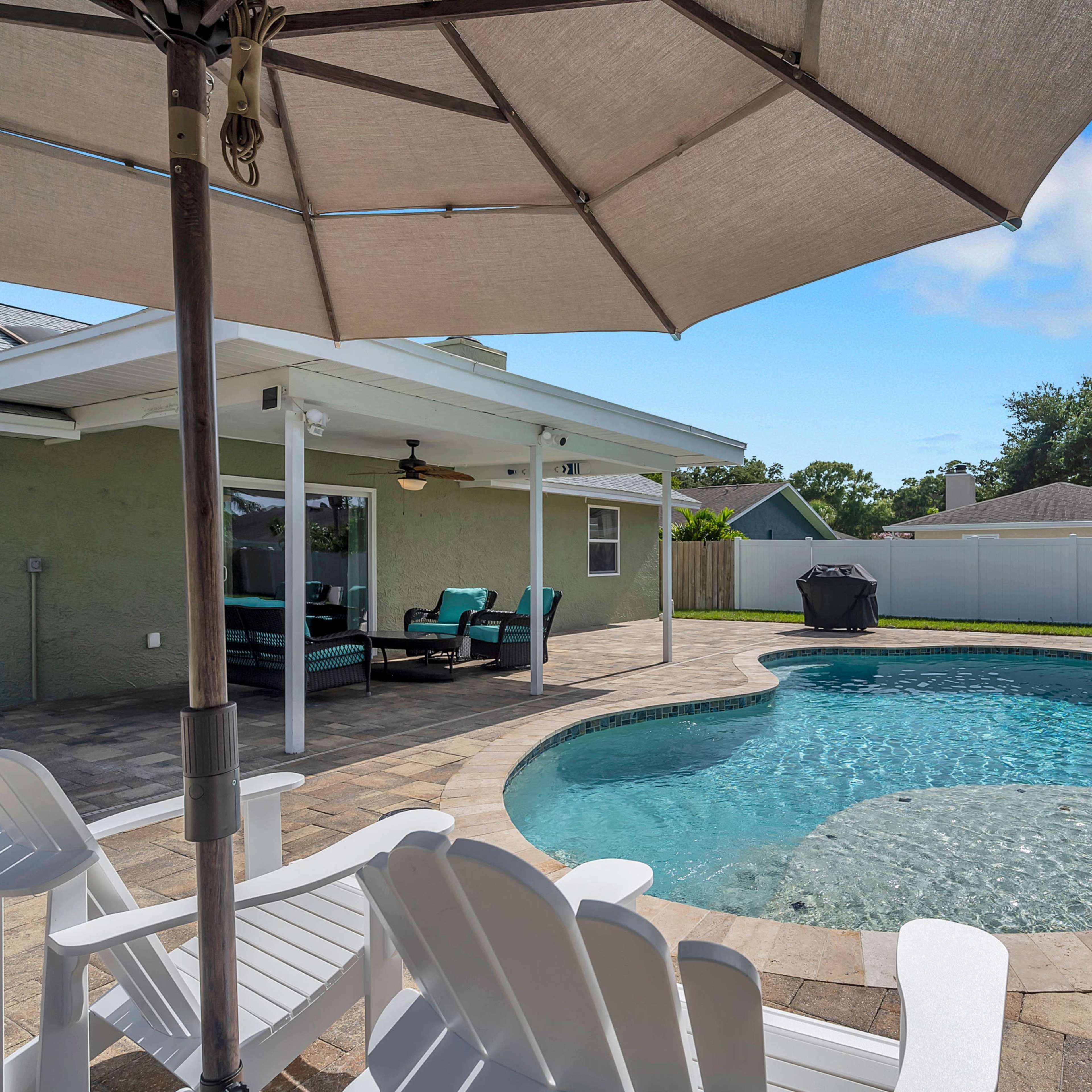 A backyard with a swimming pool surrounded by patio stones, an umbrella, and lounge chairs under a clear sky.