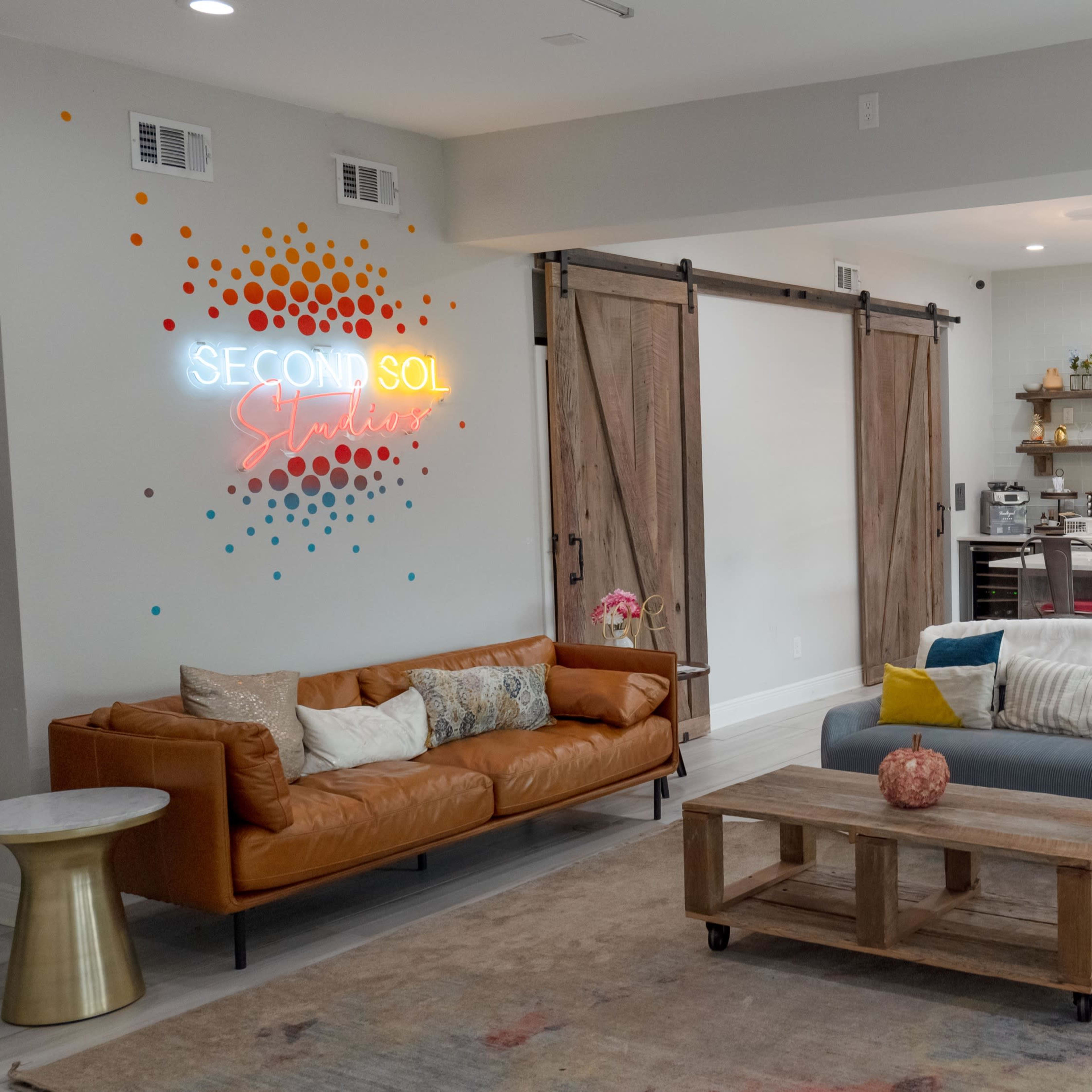 A modern living space with a brown leather sofa, a blue couch, a wooden coffee table, and a kitchen featuring red accents and hanging light fixtures.