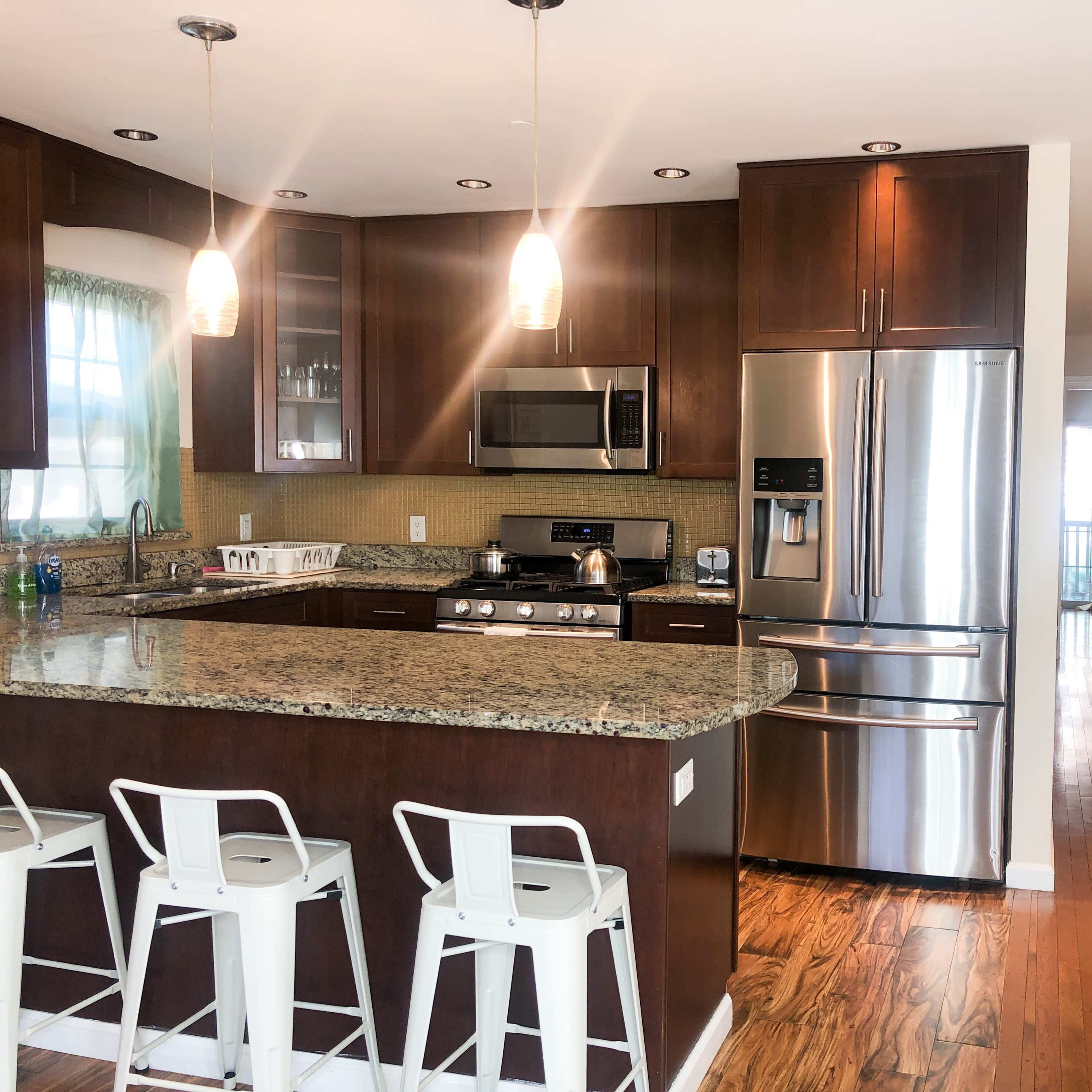 A modern kitchen with a granite countertop, stainless steel appliances, bar seating, and a view of an adjoining hallway.